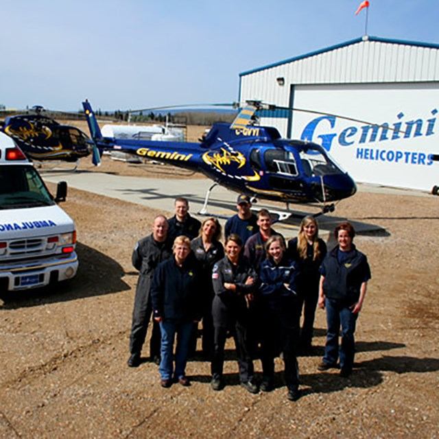 Group stands in front of Gemini helicopter outside hangar with Gemini Helicopters sign.