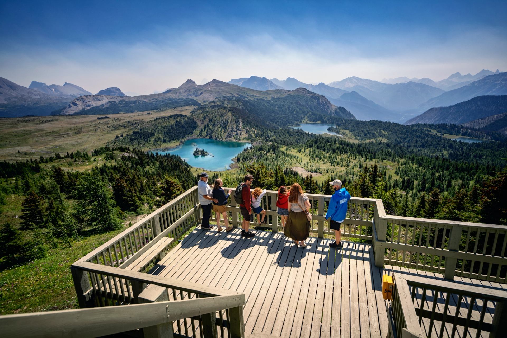 Visitors stand on a wooden lookout deck overlooking turquoise alpine lakes and mountains.