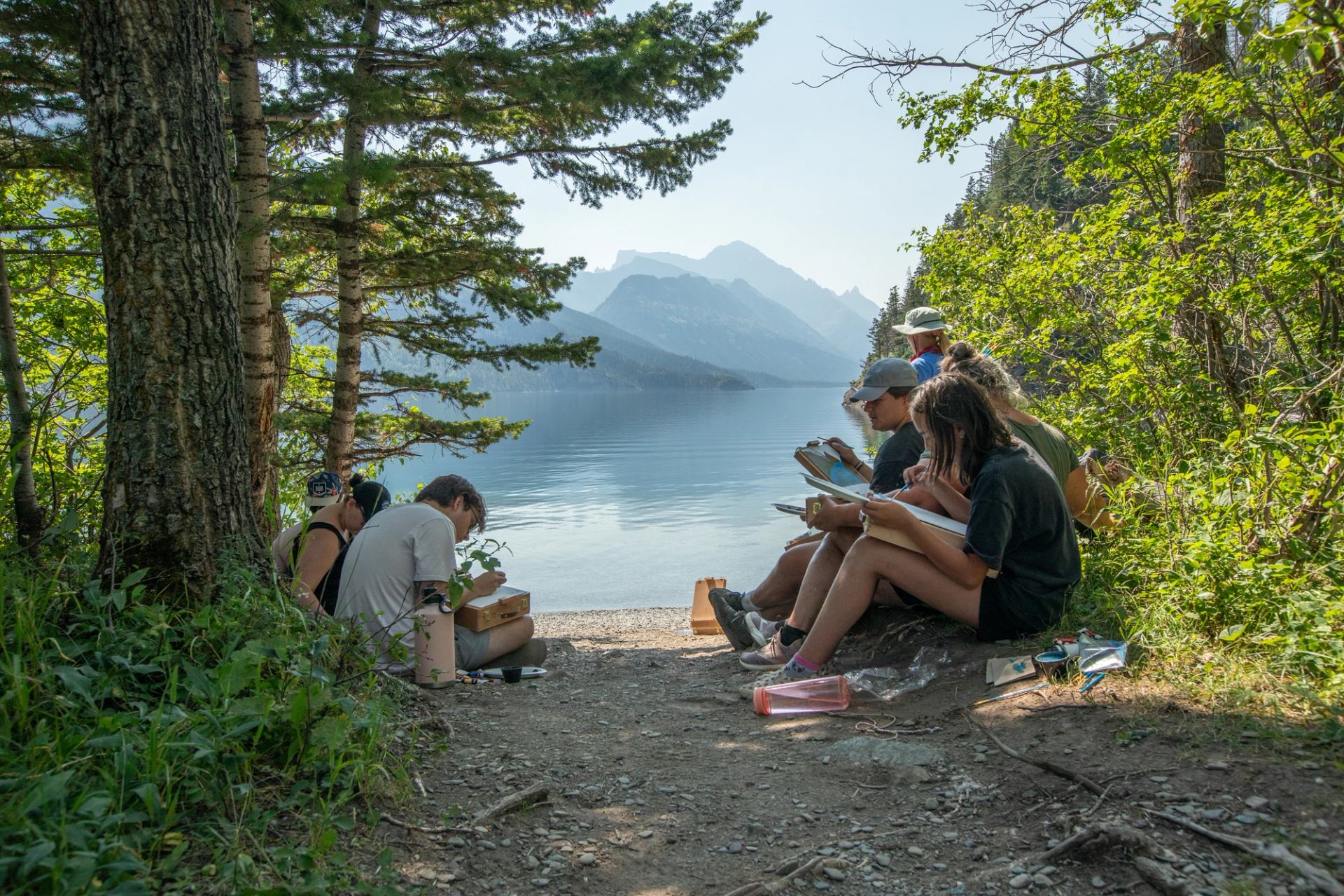 Group of people sketching beside a calm lake, surrounded by trees and mountains.