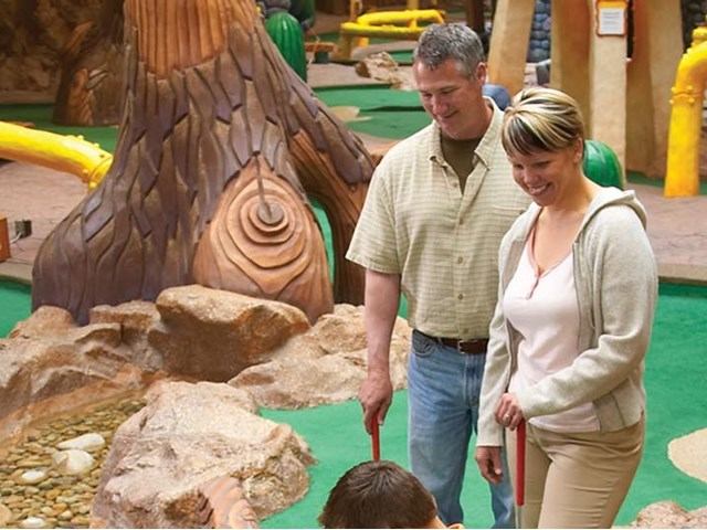 Two people hold putters beside rocks and tree sculpture on mini-golf course.