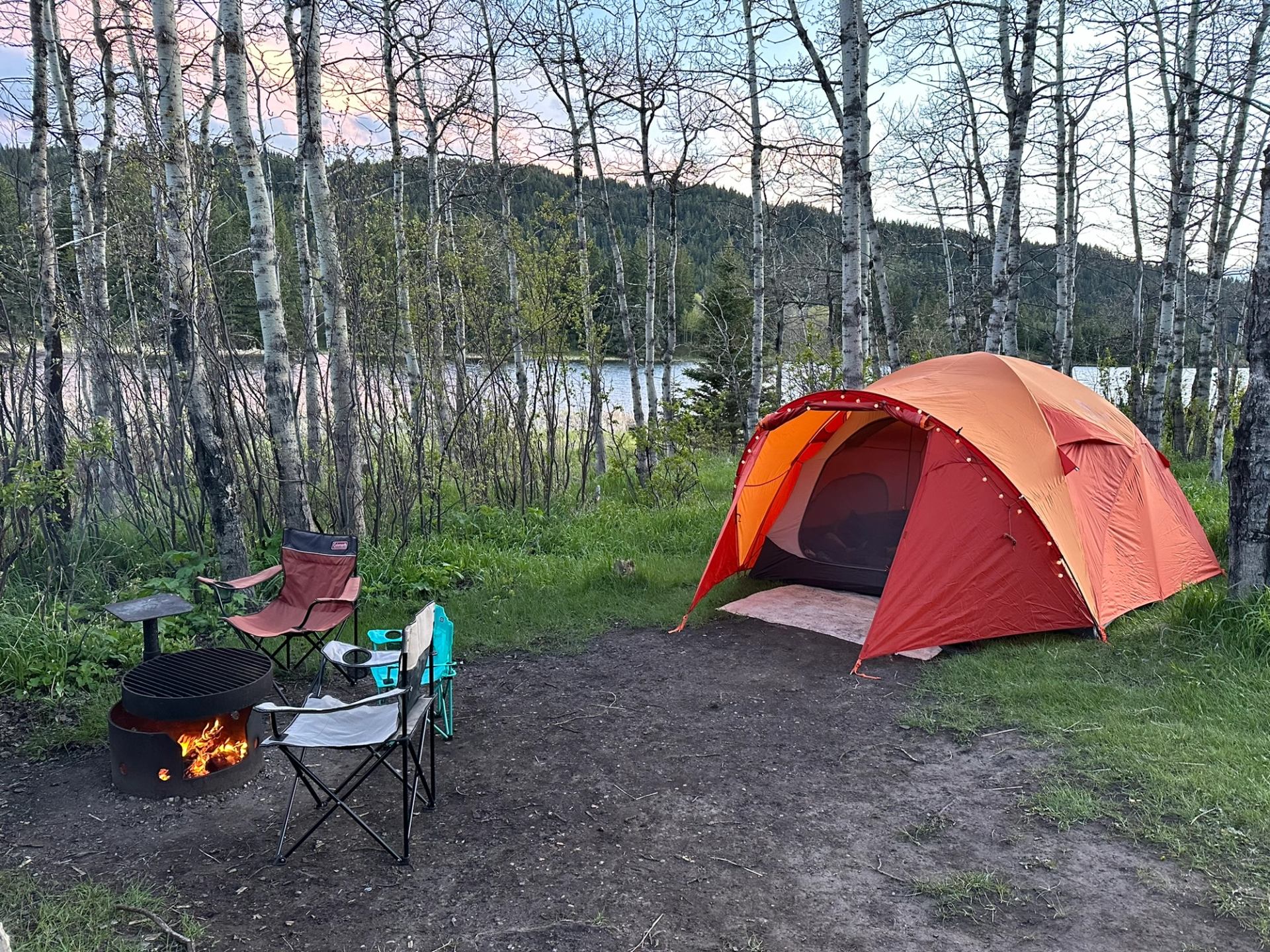 A lakeside campsite with a tent, campfire, and chairs among tall trees.