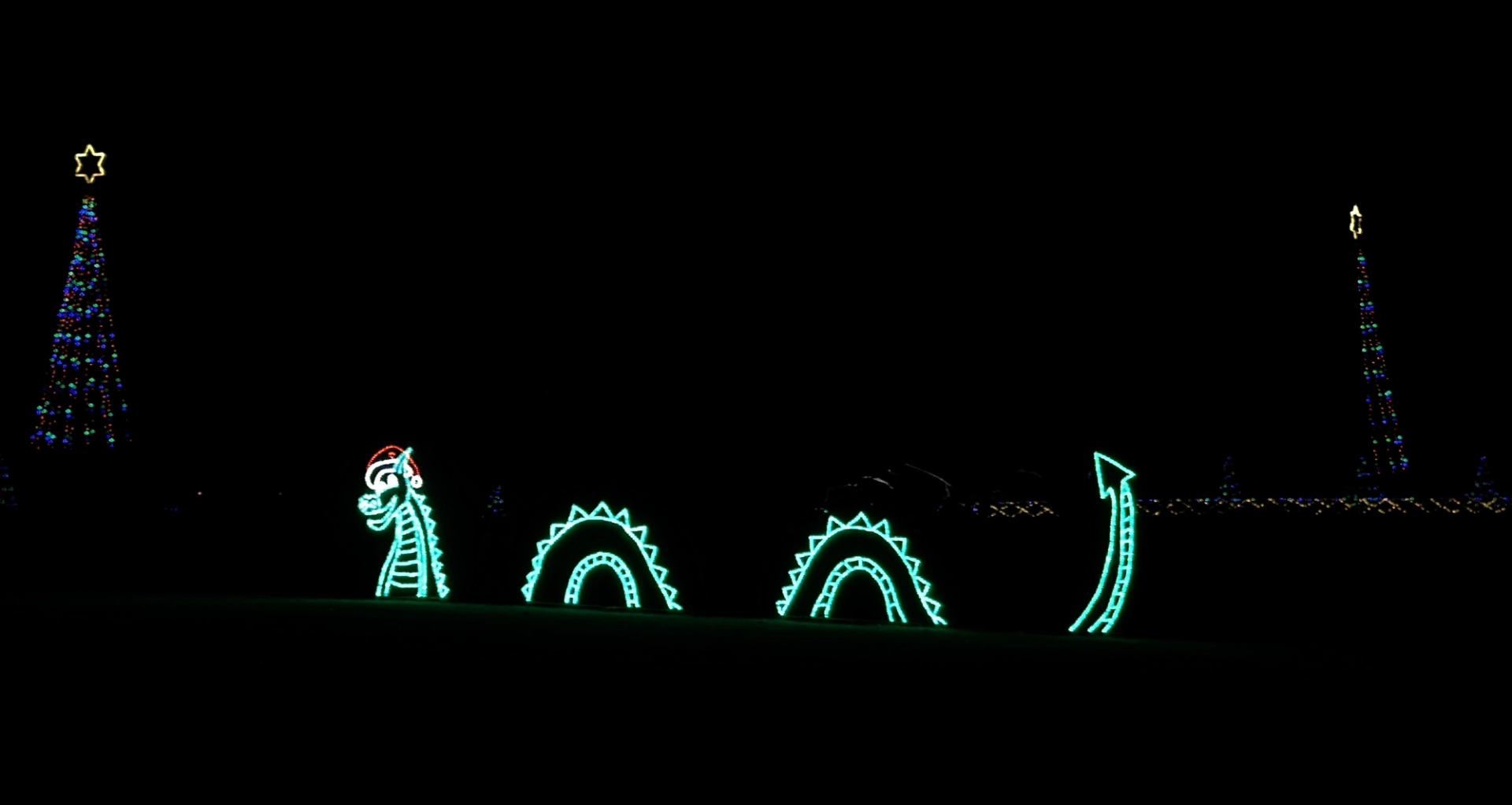 Bright green sea serpent light display with Santa hat and two lit Christmas trees nearby