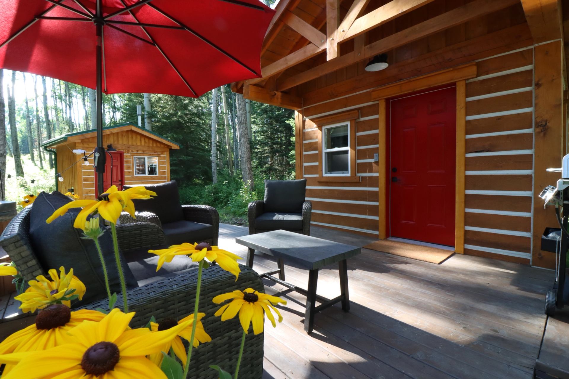 Wicker patio set under red umbrella on deck, with cabins and yellow flowers in forest setting.