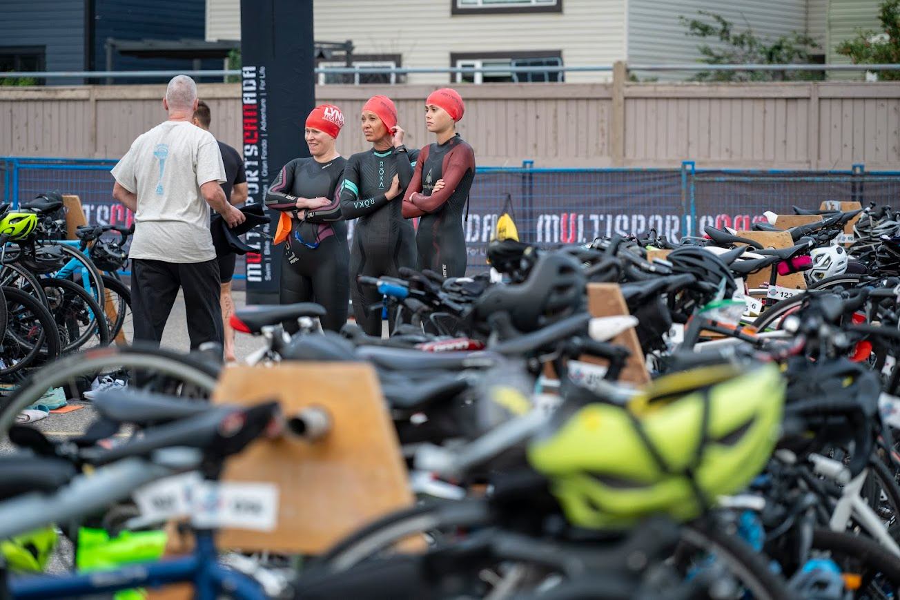 Triathletes in wetsuits and red swim caps stand near the bike transition area at Lake Chaparral Triathlon.