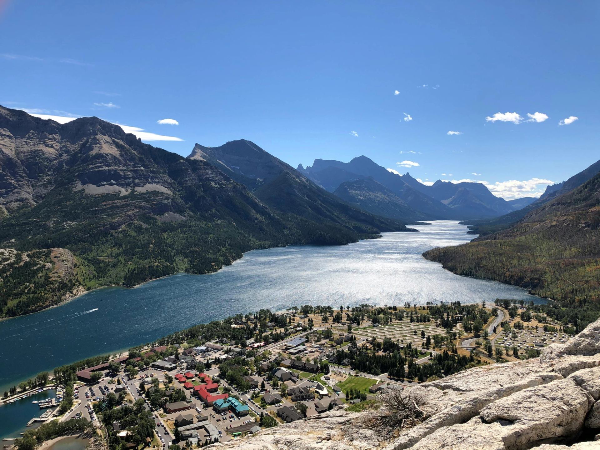 Mountain lake valley with town, marina and winding road under clear blue sky.