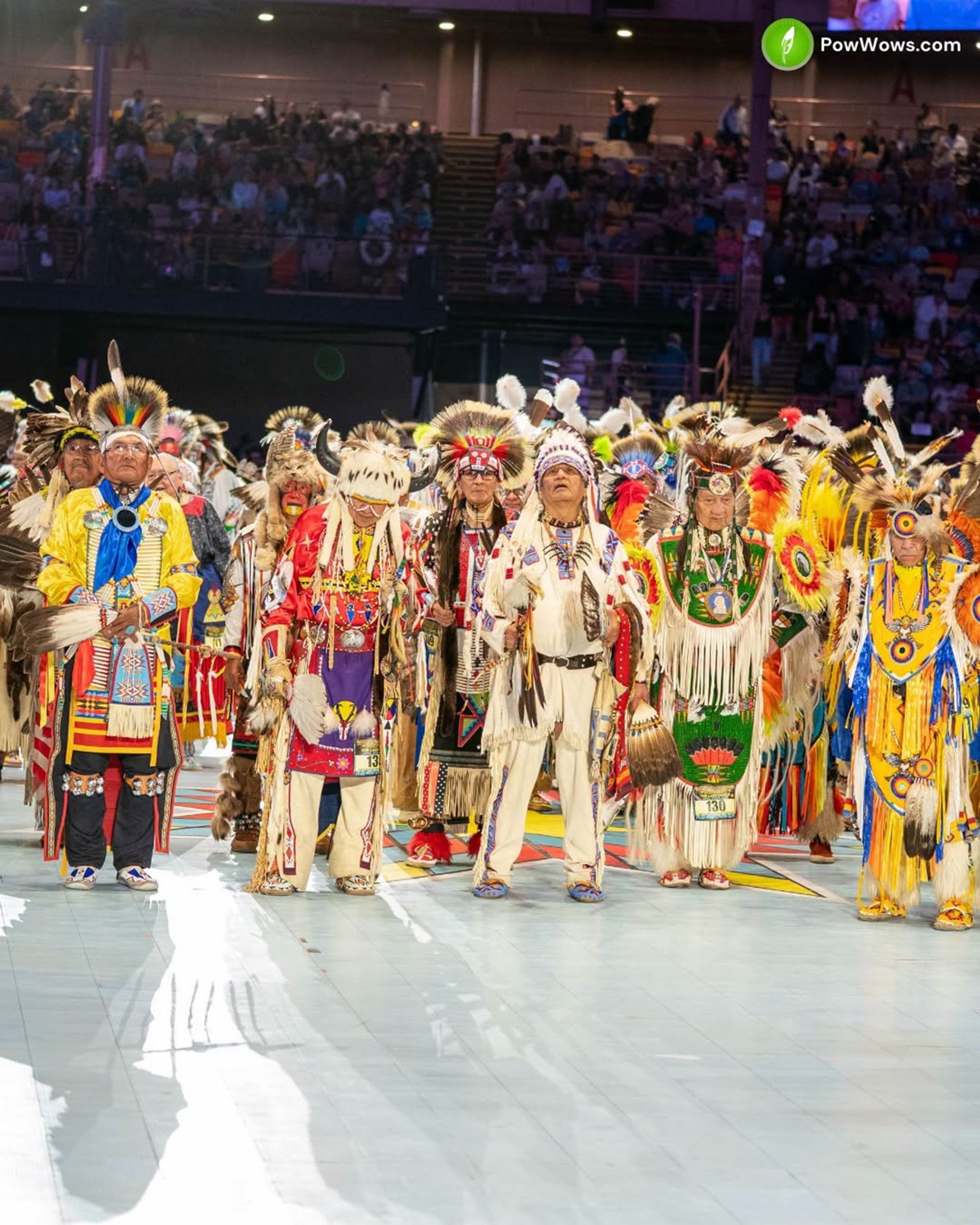 Indigenous dancers in colourful regalia stand in an arena during a powwow event.