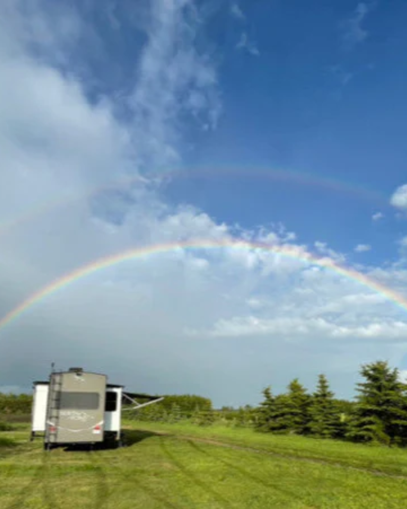 Double rainbow arching over RV parked on grassy field beside evergreen trees.