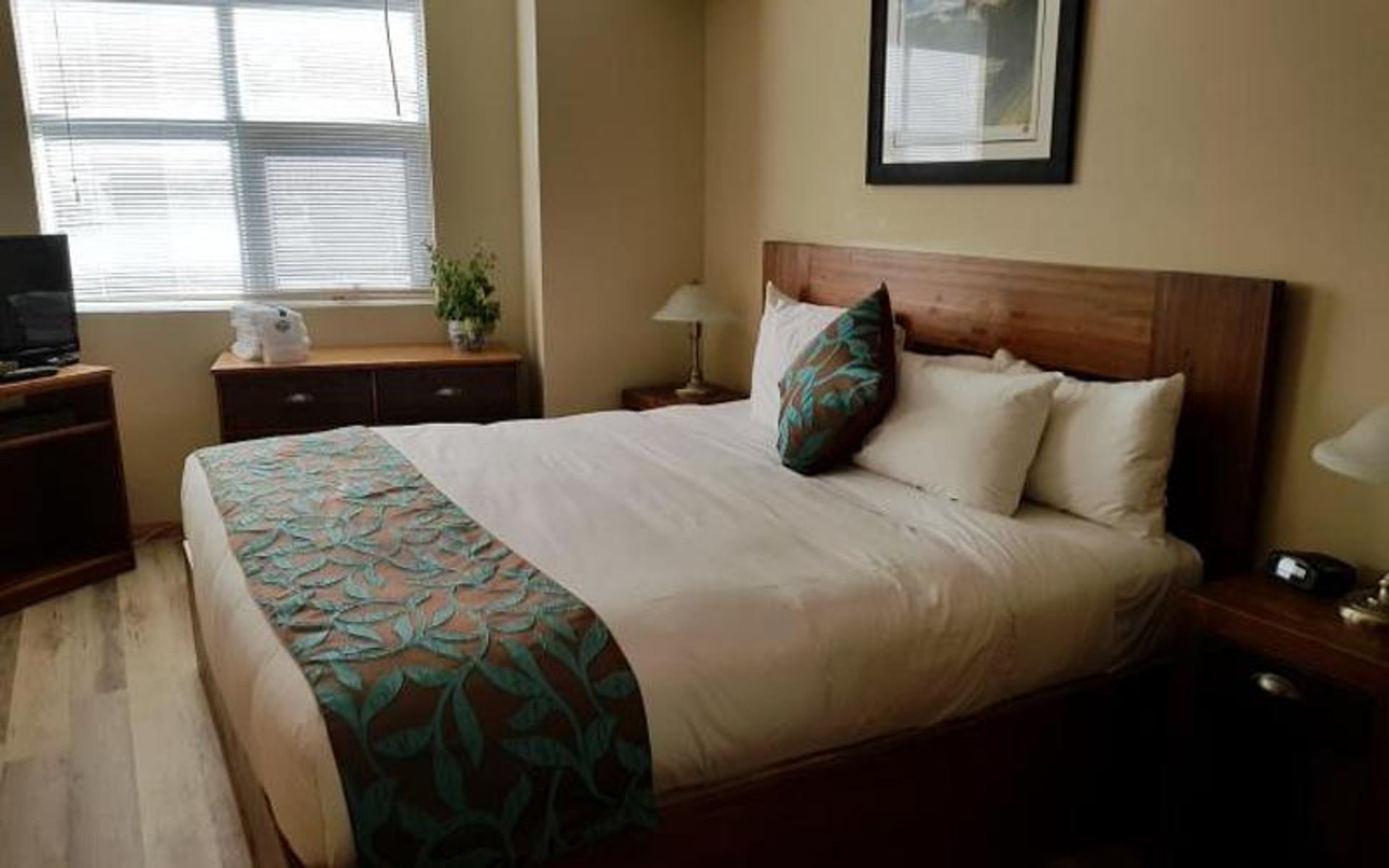 Hotel-style bedroom with double bed, patterned runner, wooden furniture and window blinds.