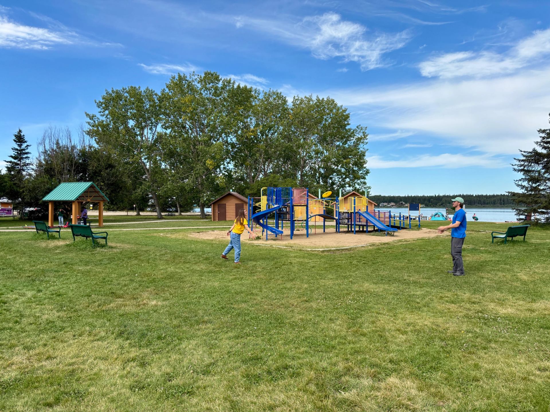 People playing near colourful playground equipment beside a lake, with trees and benches.