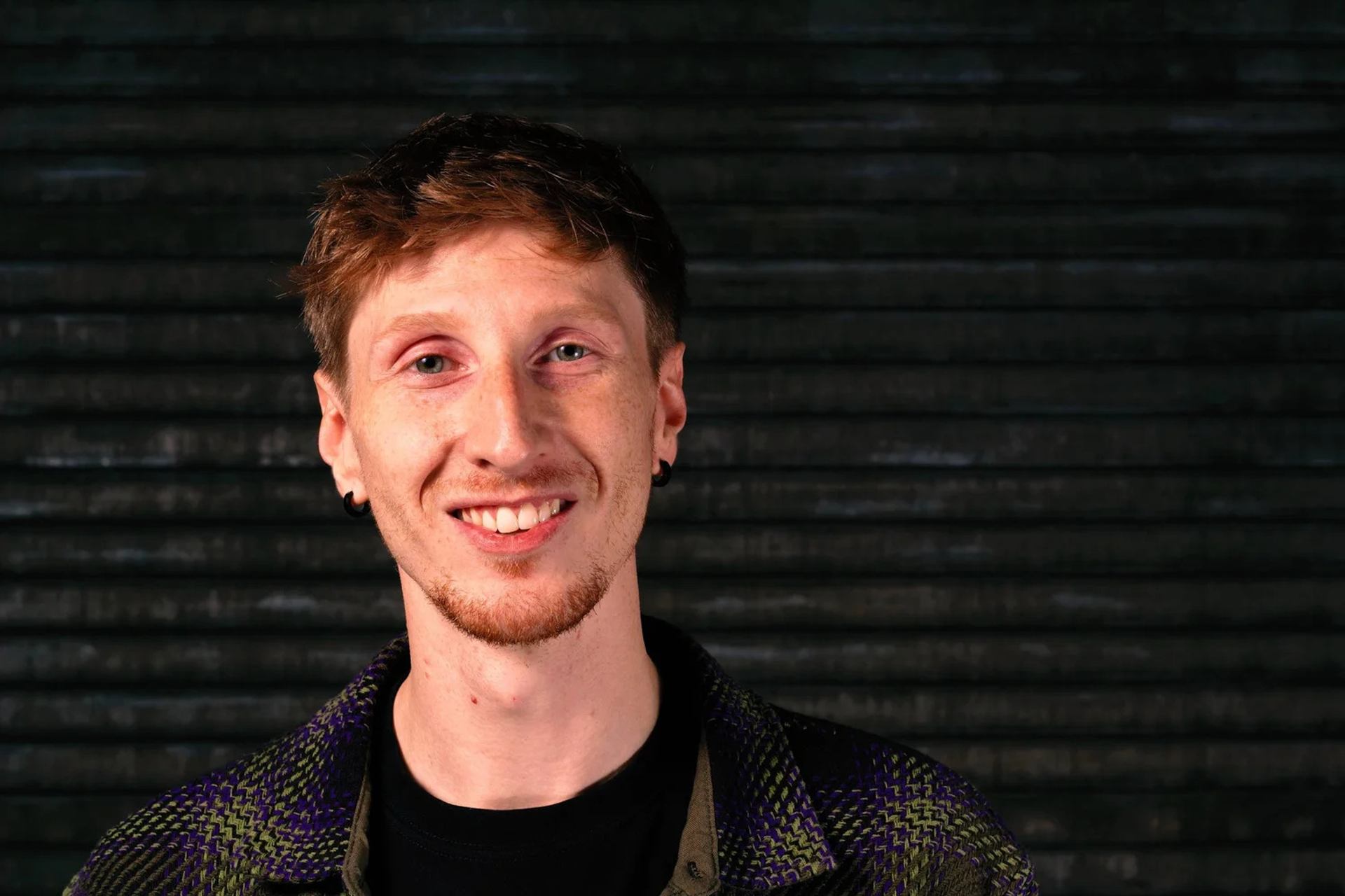 Person with short hair and earrings standing before dark metal shutter background.