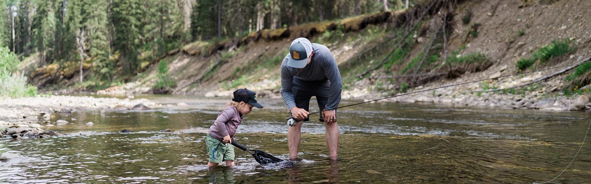 Man and child stand in shallow creek, fishing with net and rod amid trees