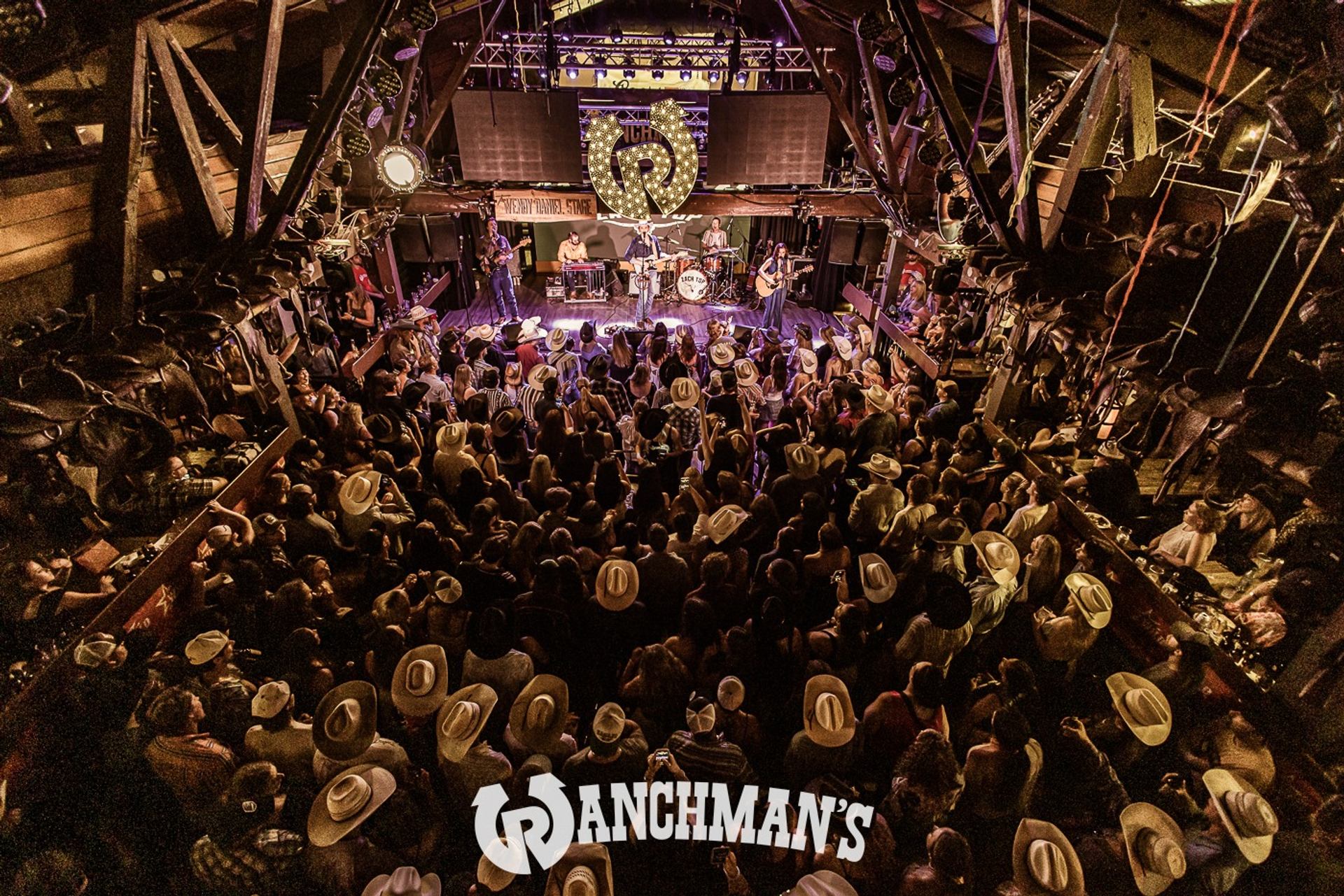 Crowded Ranchman’s bar with cowboy hats watching live band perform on indoor stage.