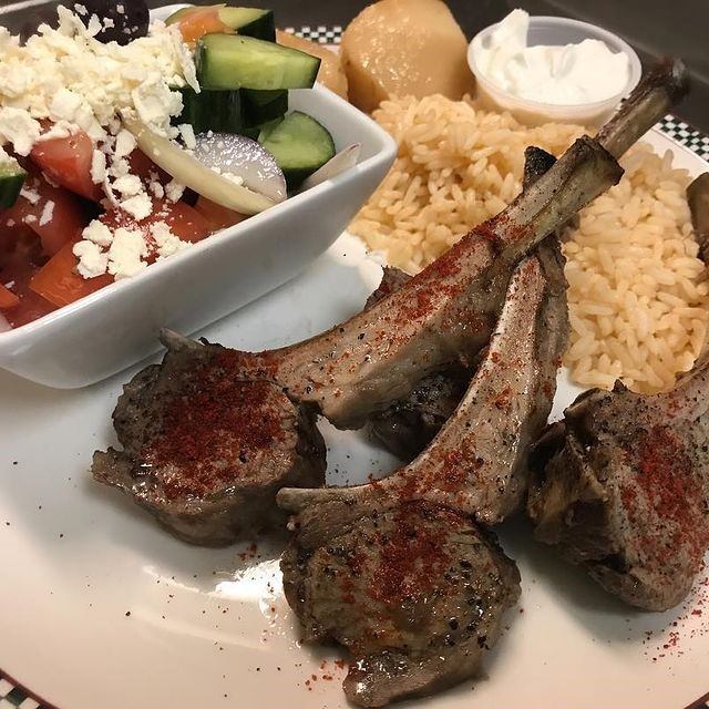 Plate with grilled lamb chops, rice, Greek salad, and white sauce cup.