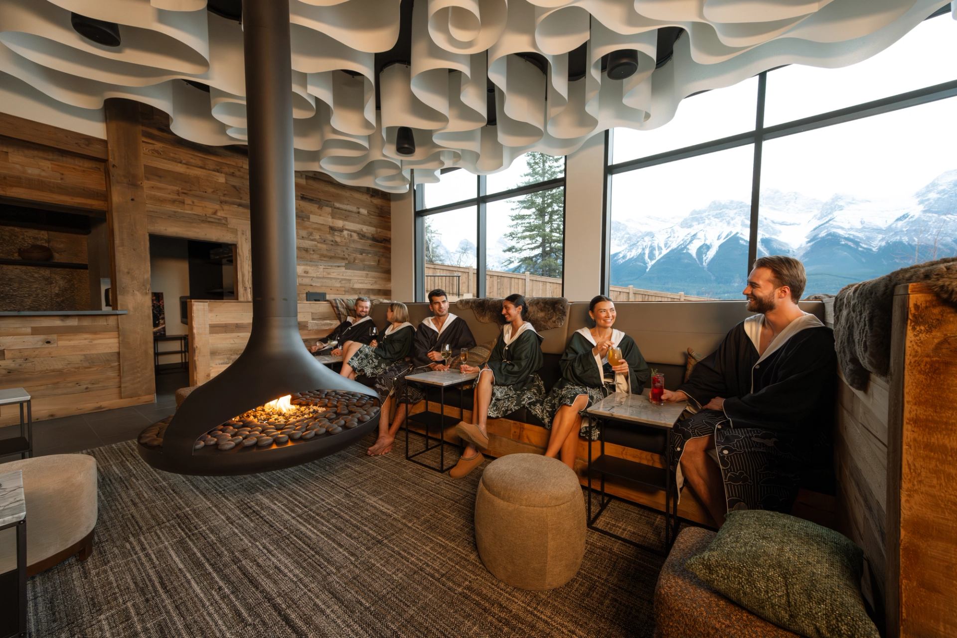 Guests relaxing by a modern fireplace in Everwild Canmore Nordic Spa lounge with mountain views.