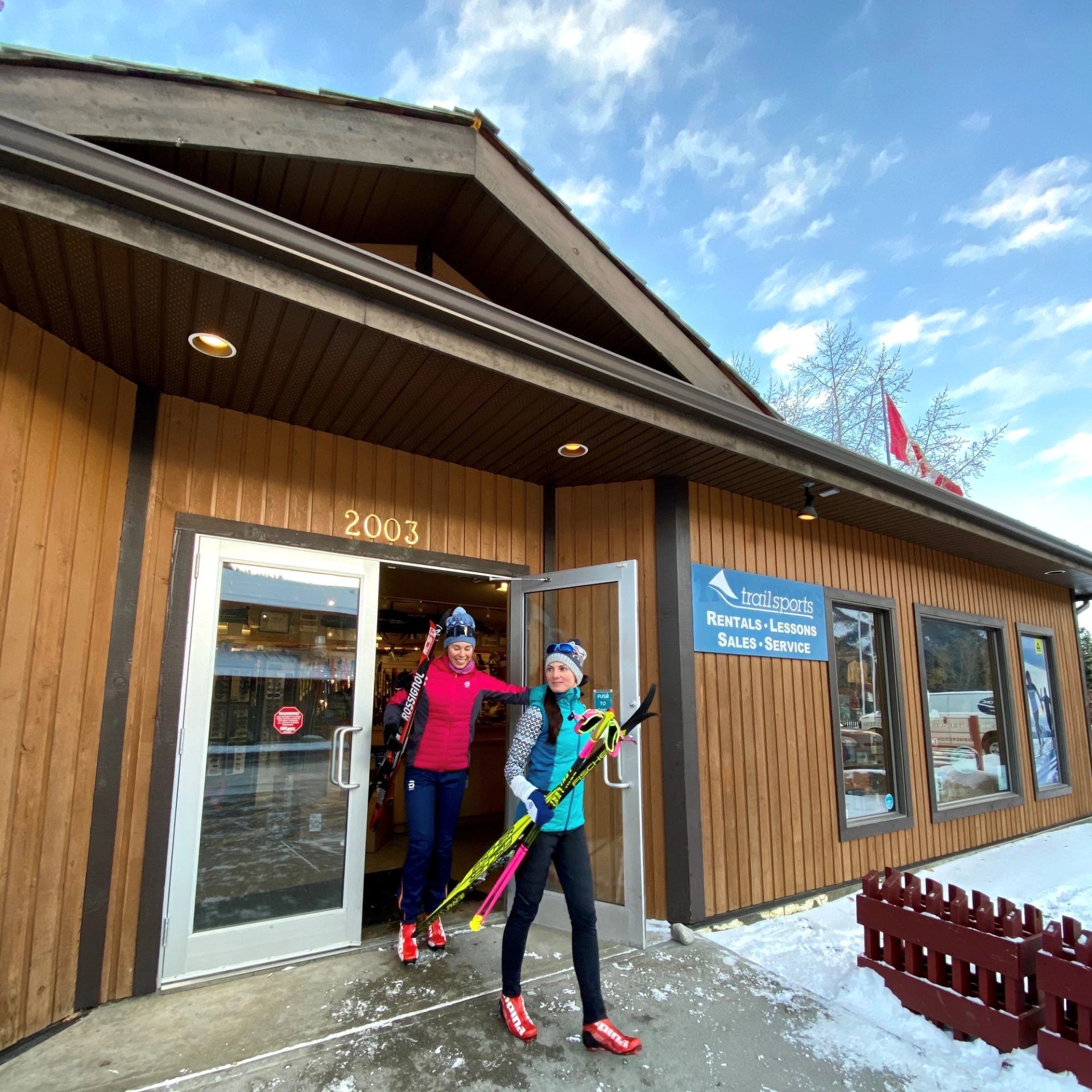 Two women in winter gear exit the shop with cross-country skis on a snowy day.