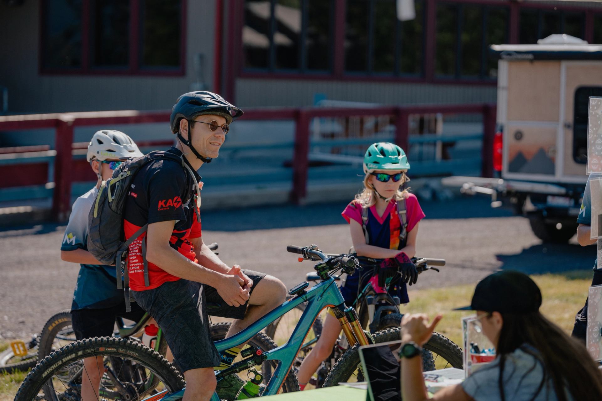 Cyclists wearing helmets stand with mountain bikes near an outdoor registration or information table.
