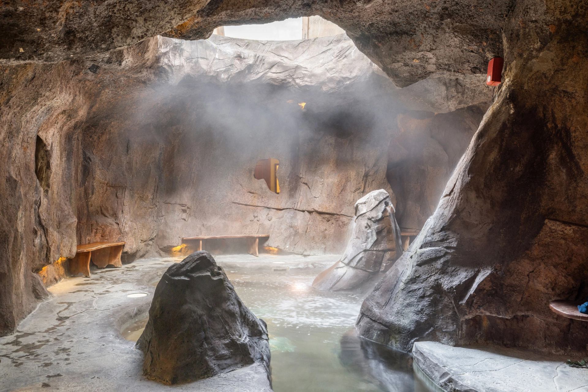 Rocky indoor grotto with steaming pool, stone walls and wooden benches along edges.