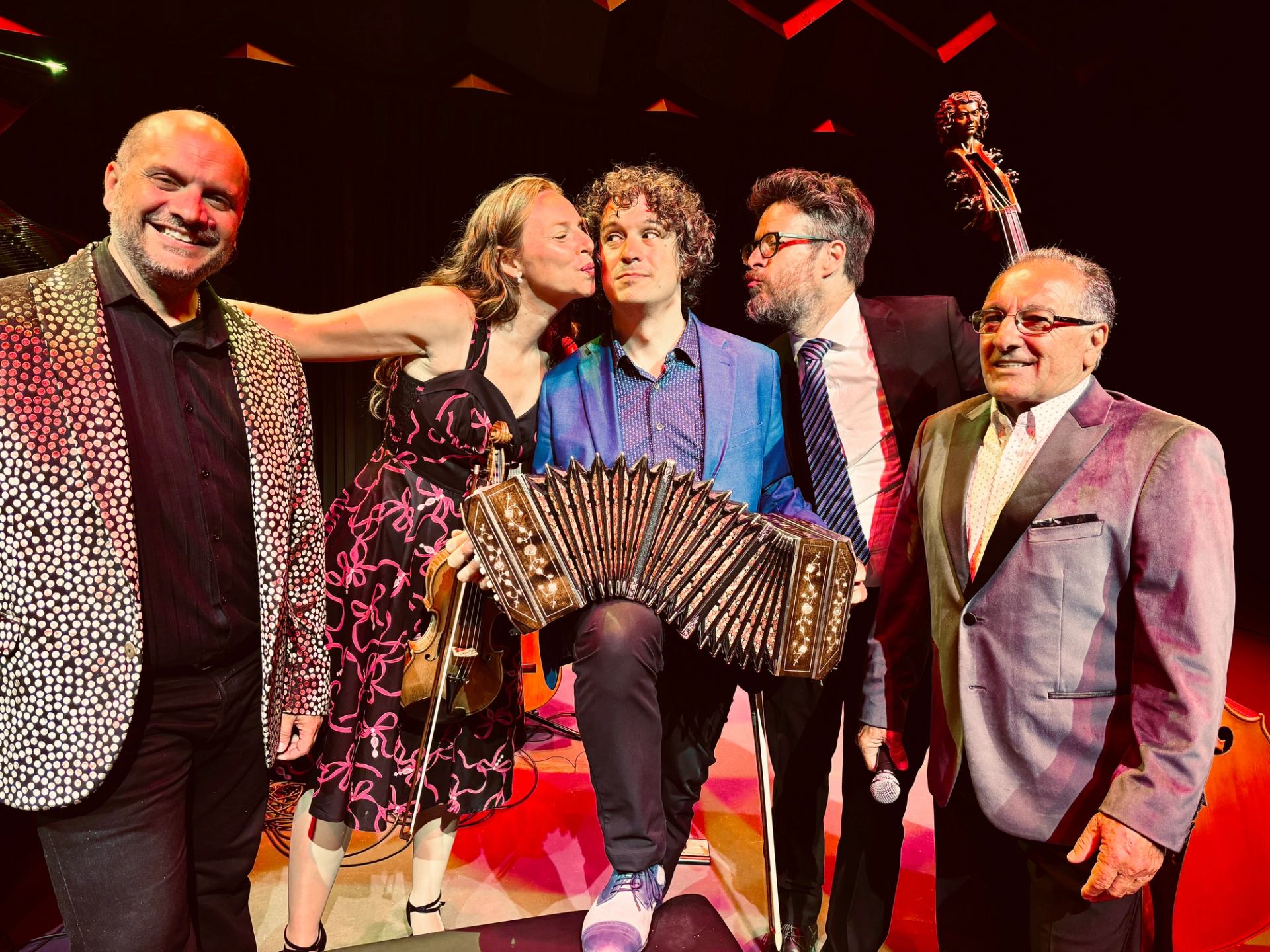 Musicians on stage pose together, central performer holding bandoneon, others smiling nearby.
