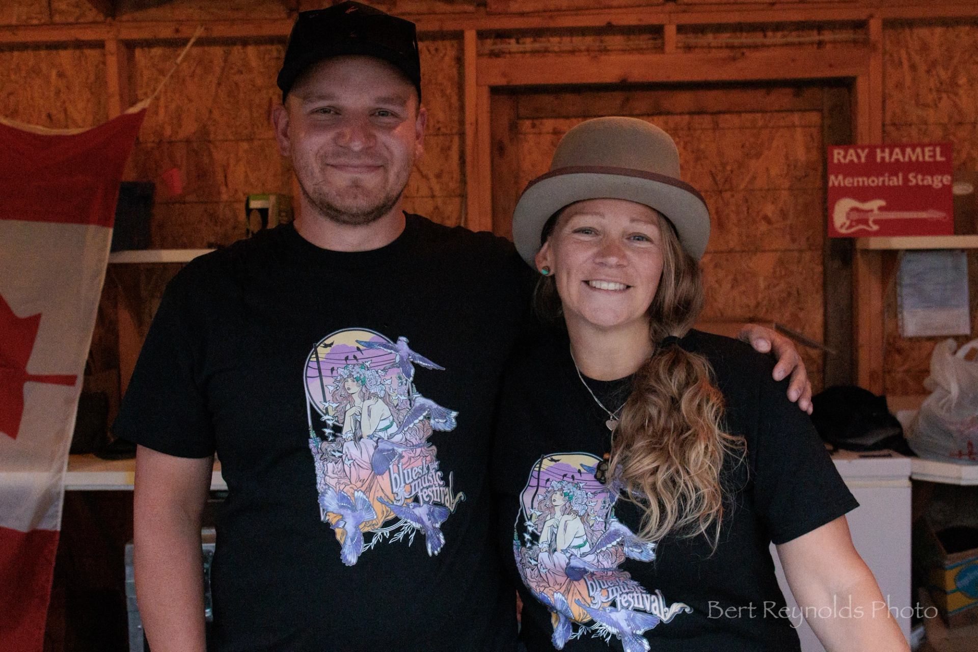 Two people in matching blues festival shirts stand indoors beside a Canadian flag.