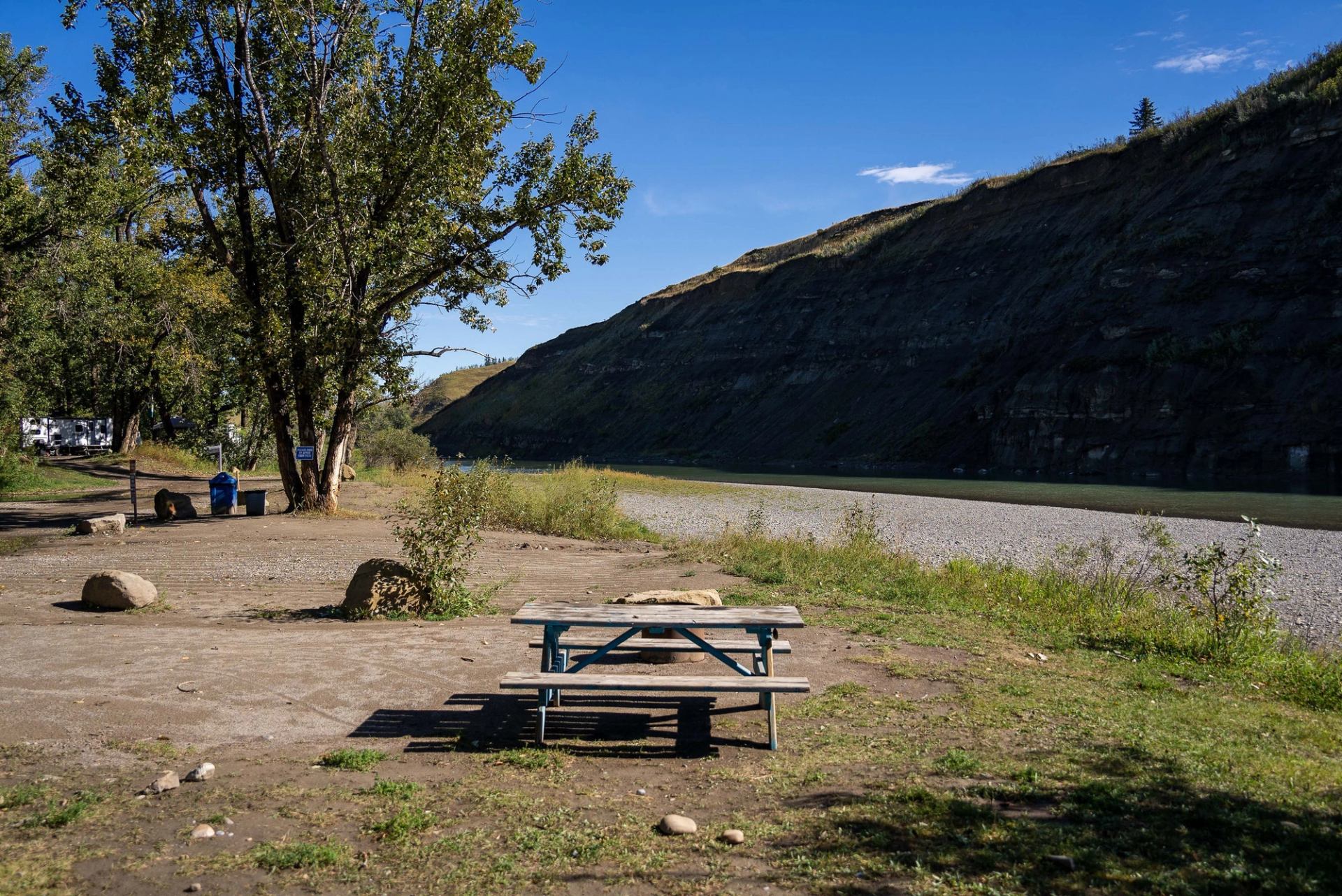 Picnic table by a river with trees and a rocky hillside in the background.