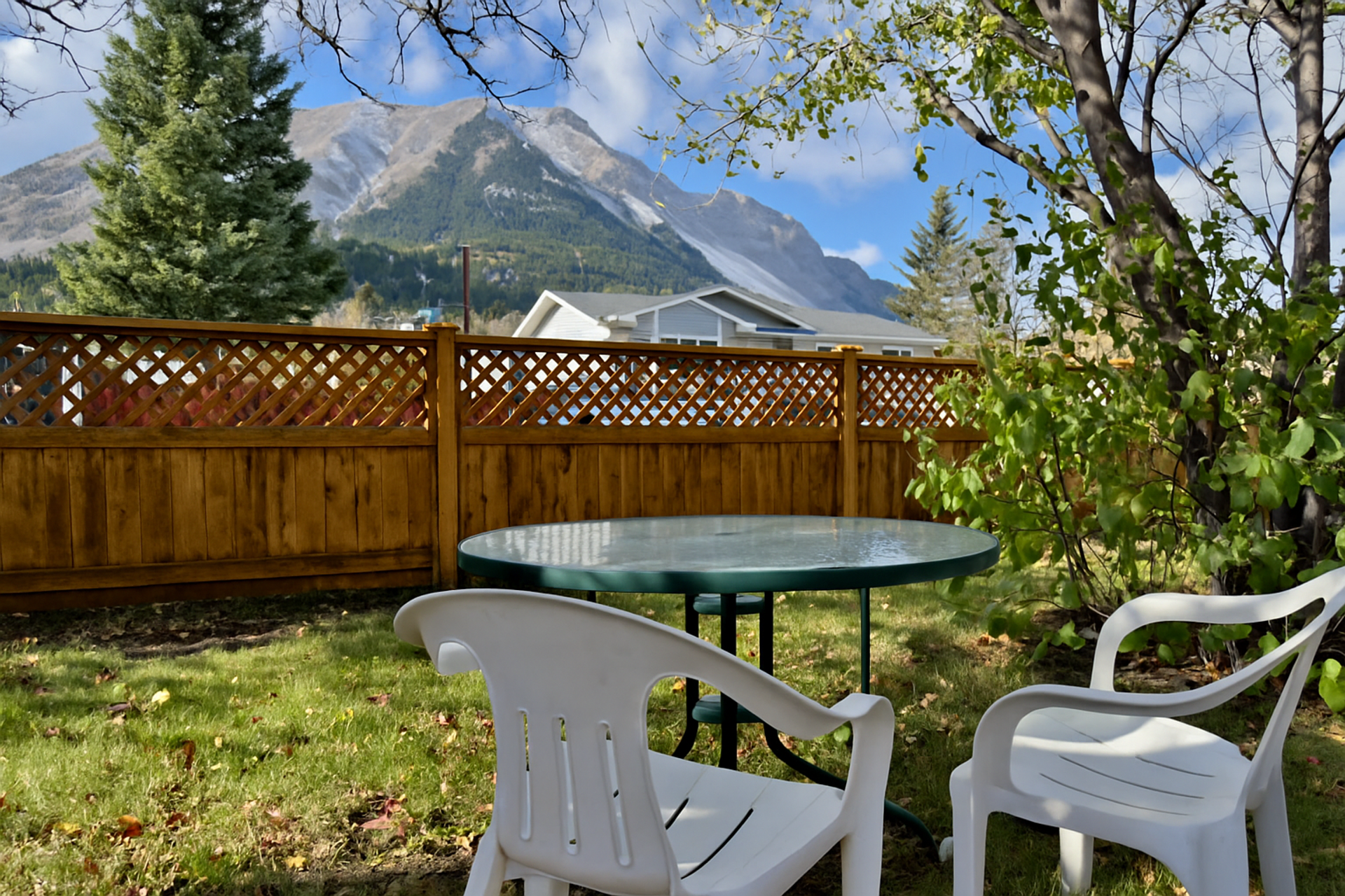 Sunny backyard with white chairs, a glass table, a wooden fence, and mountains in the background.