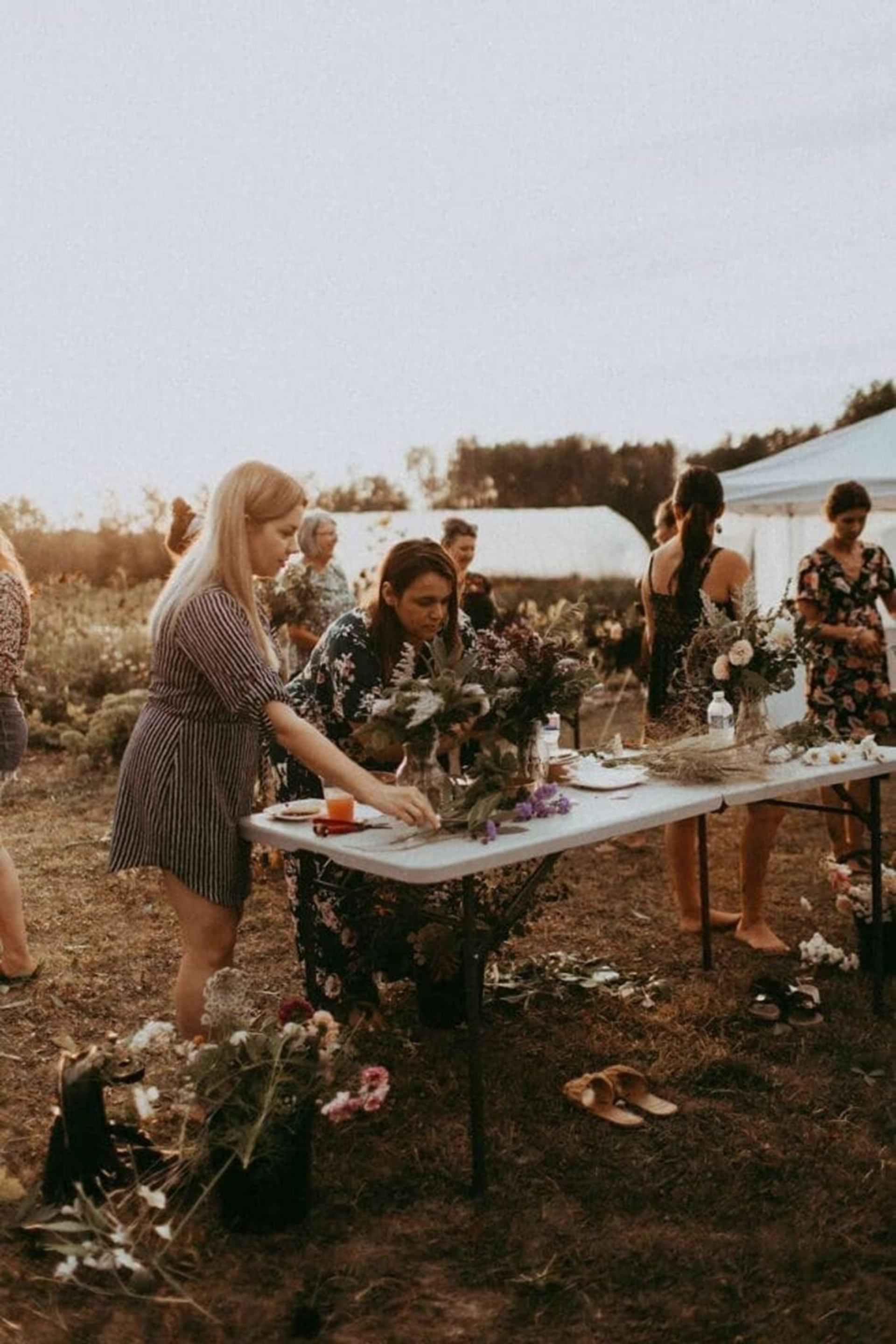 People arrange flower bouquets on folding tables in an outdoor field gathering.