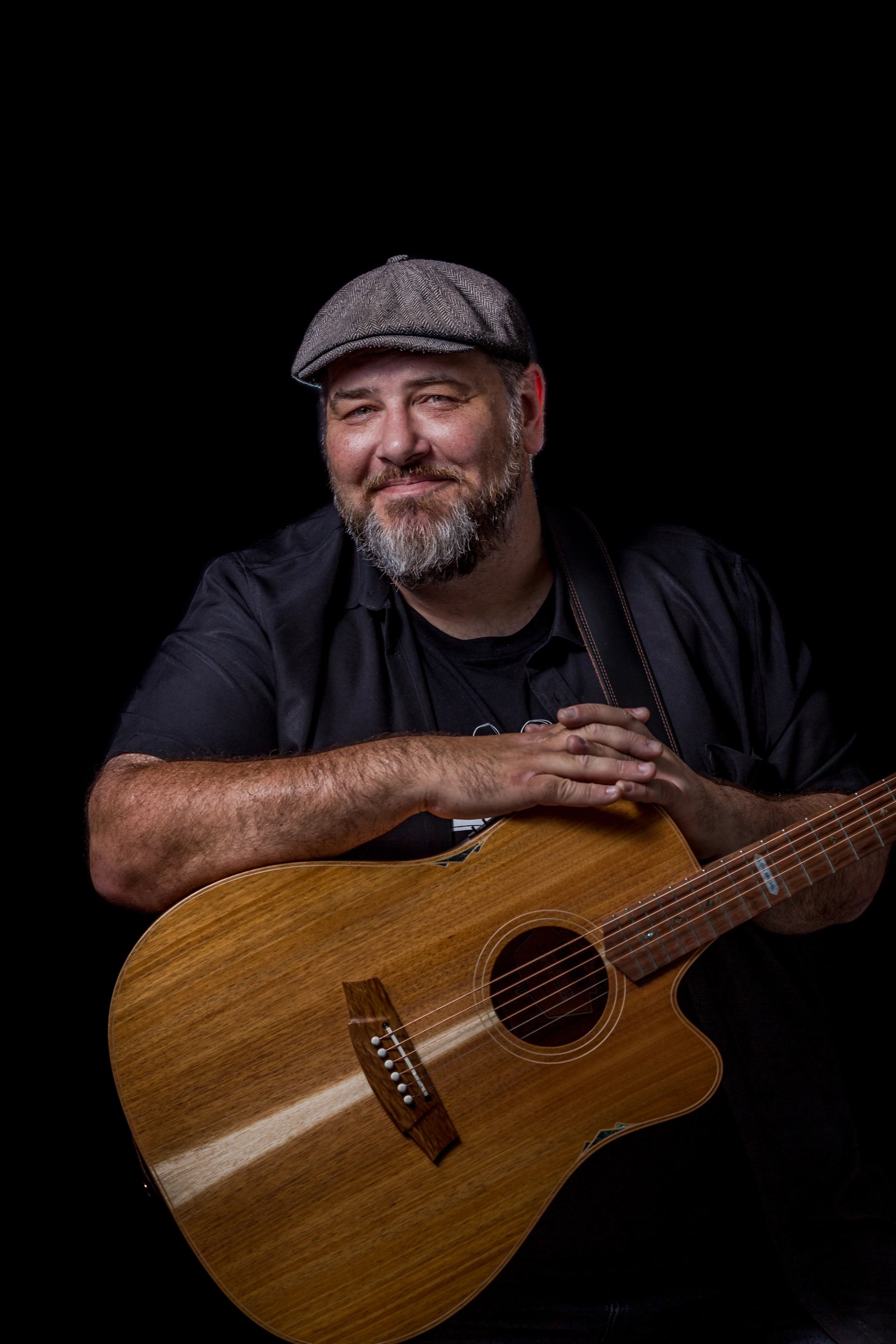 Bearded person in flat cap holding an acoustic guitar against a black background.