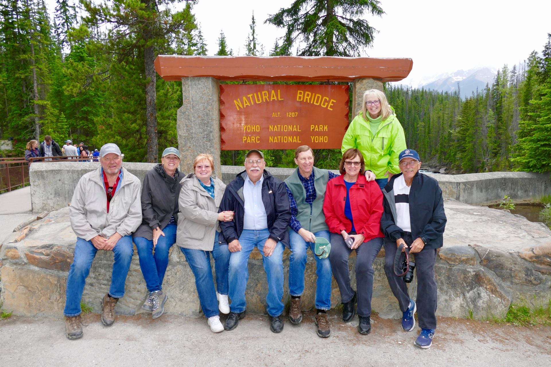 Group of people posing in front of the Natural Bridge sign at Yoho National Park, surrounded by forested mountains.