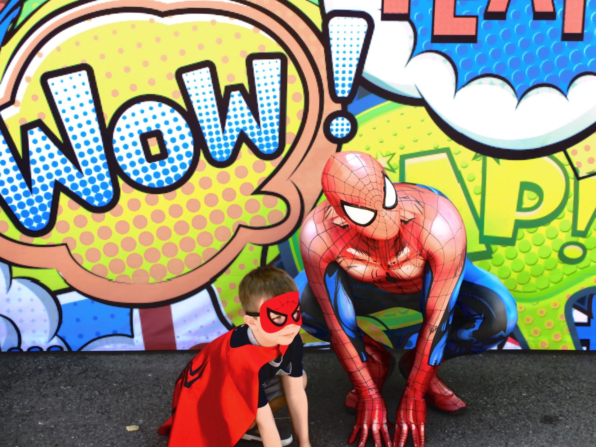 Child in cape crouches beside Spider-Man in front of comic style Wow backdrop.