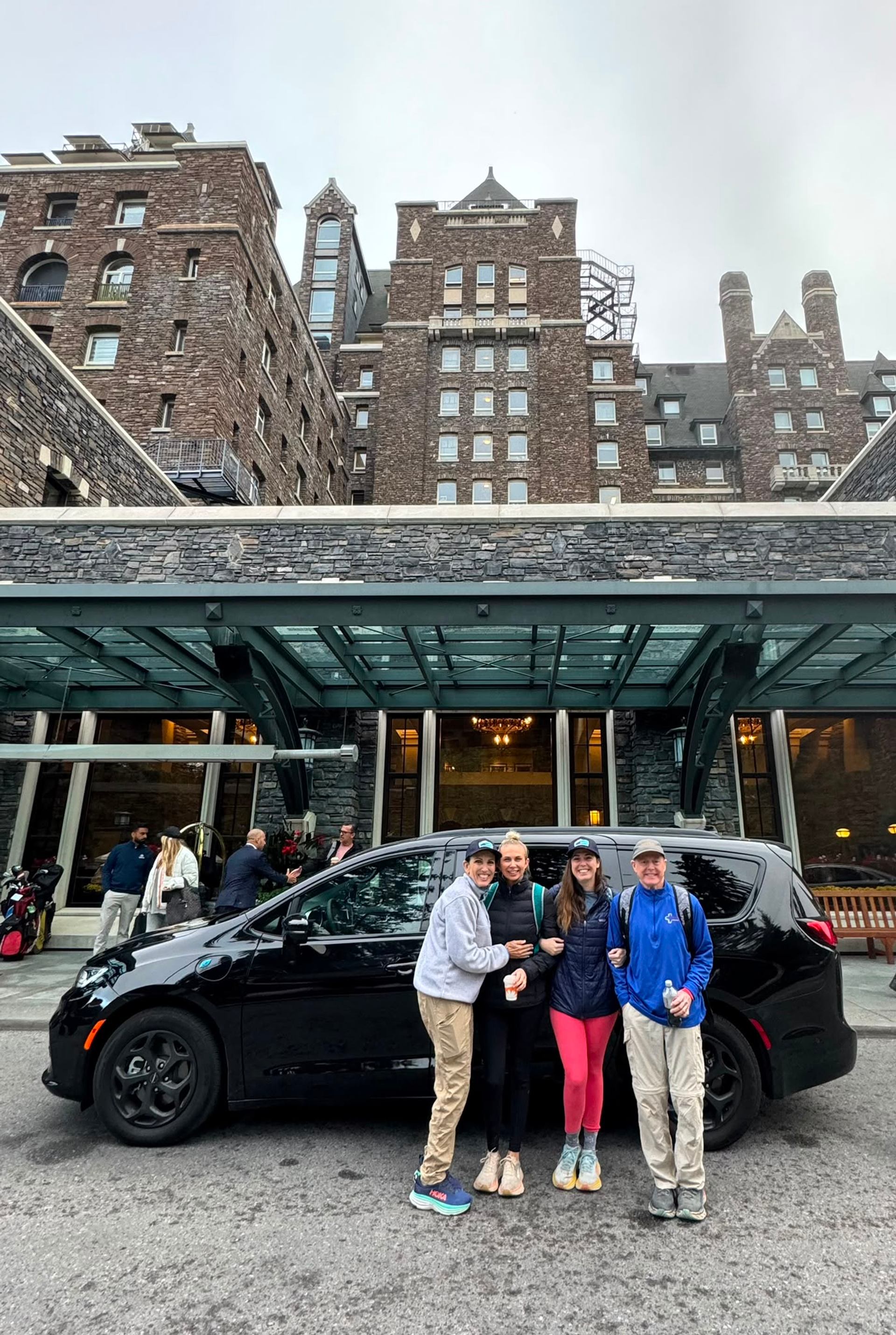 Four people stand beside a black van outside a large stone building.