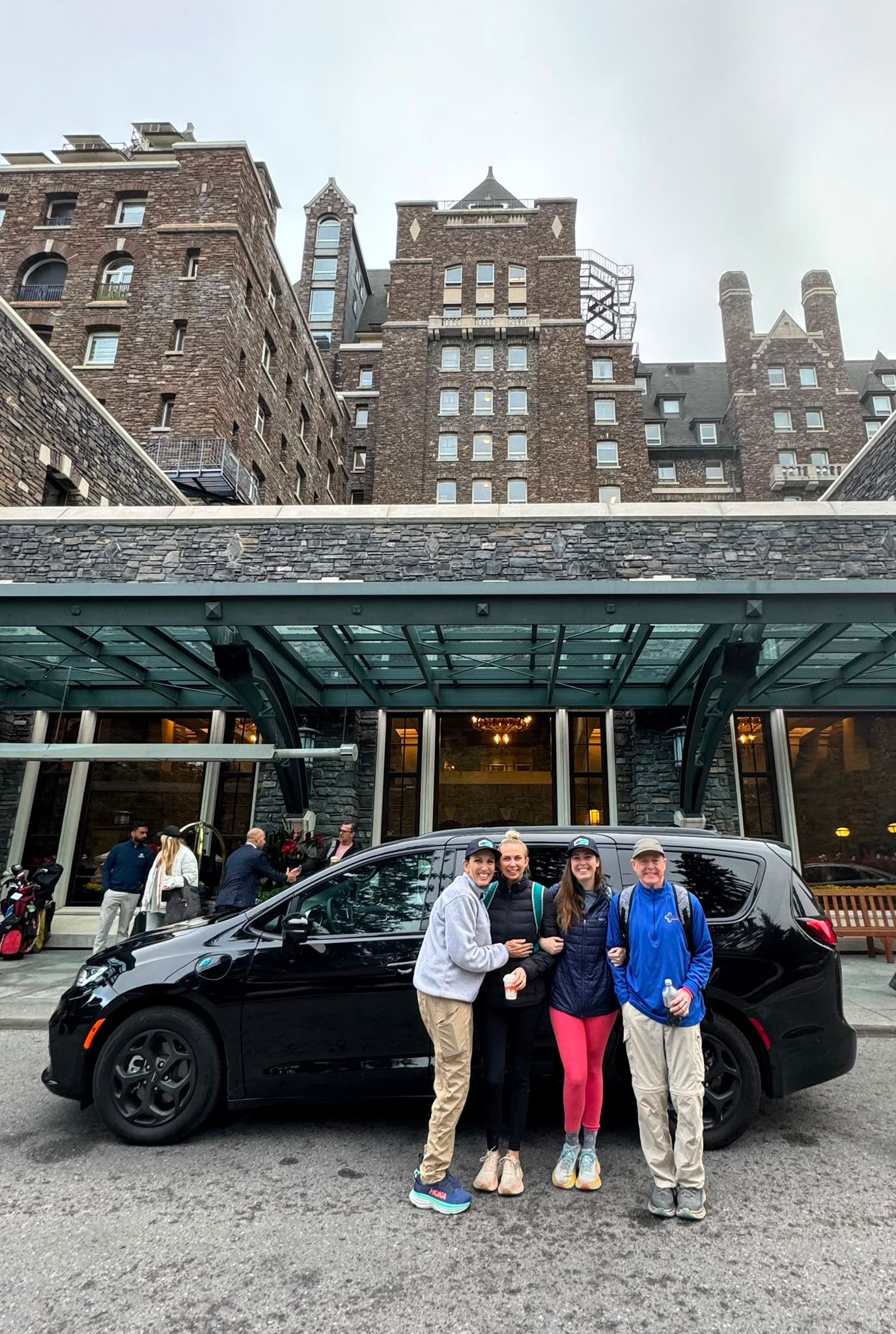 Four people stand beside a black van outside a large stone building.