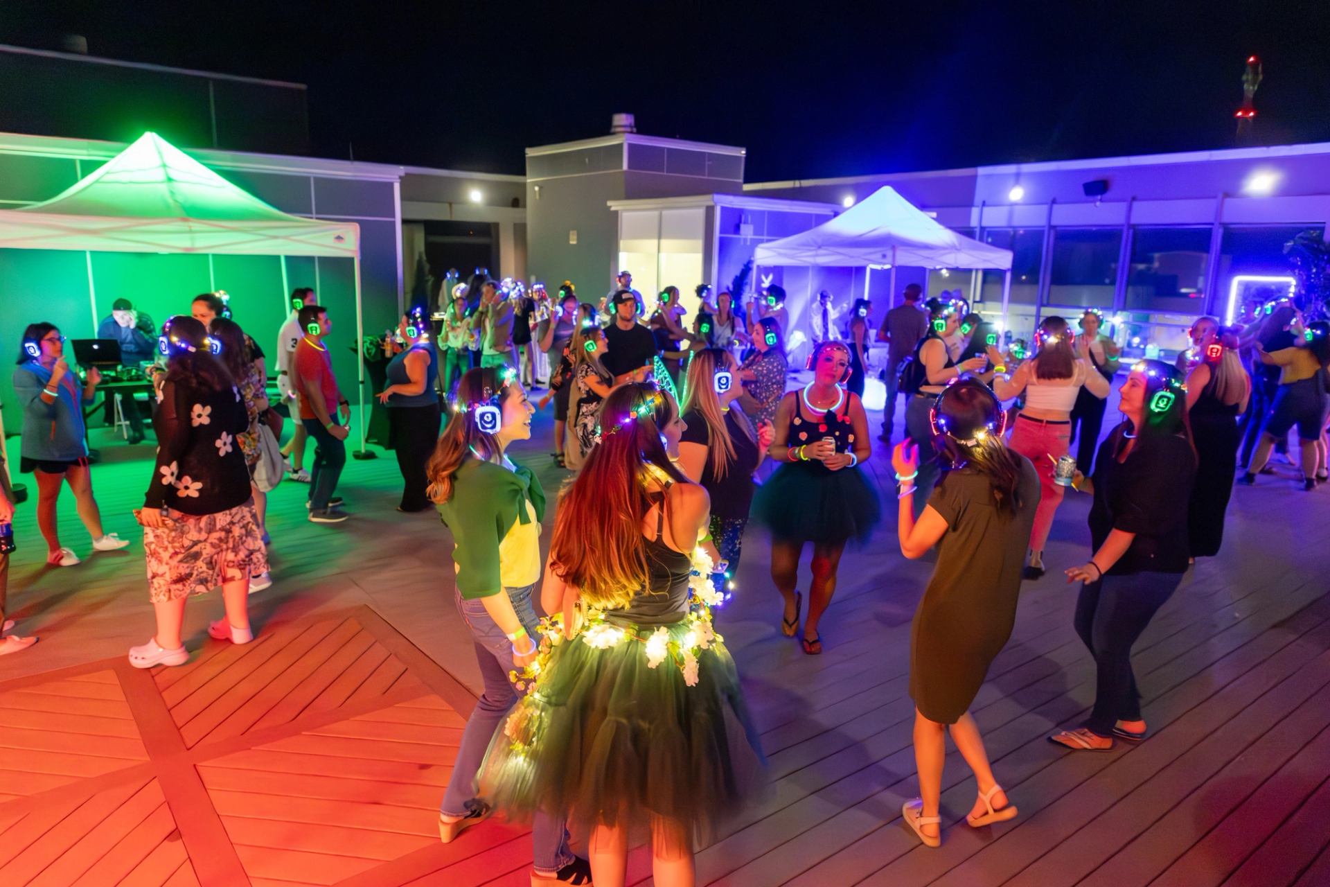 Crowd wearing glowing headphones dances on rooftop under colourful lights and white tents.