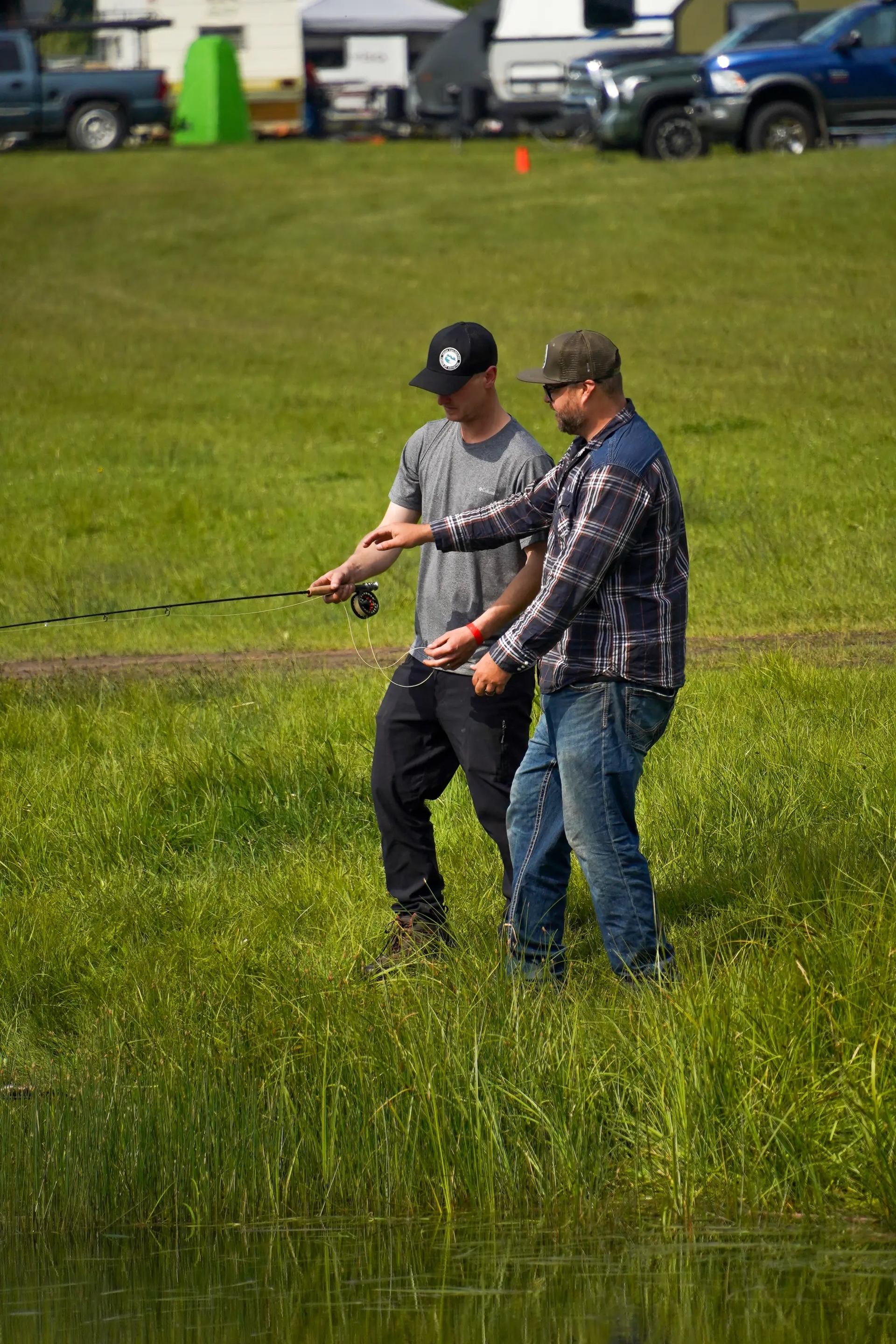 Two men fishing by a pond, one instructing the other.