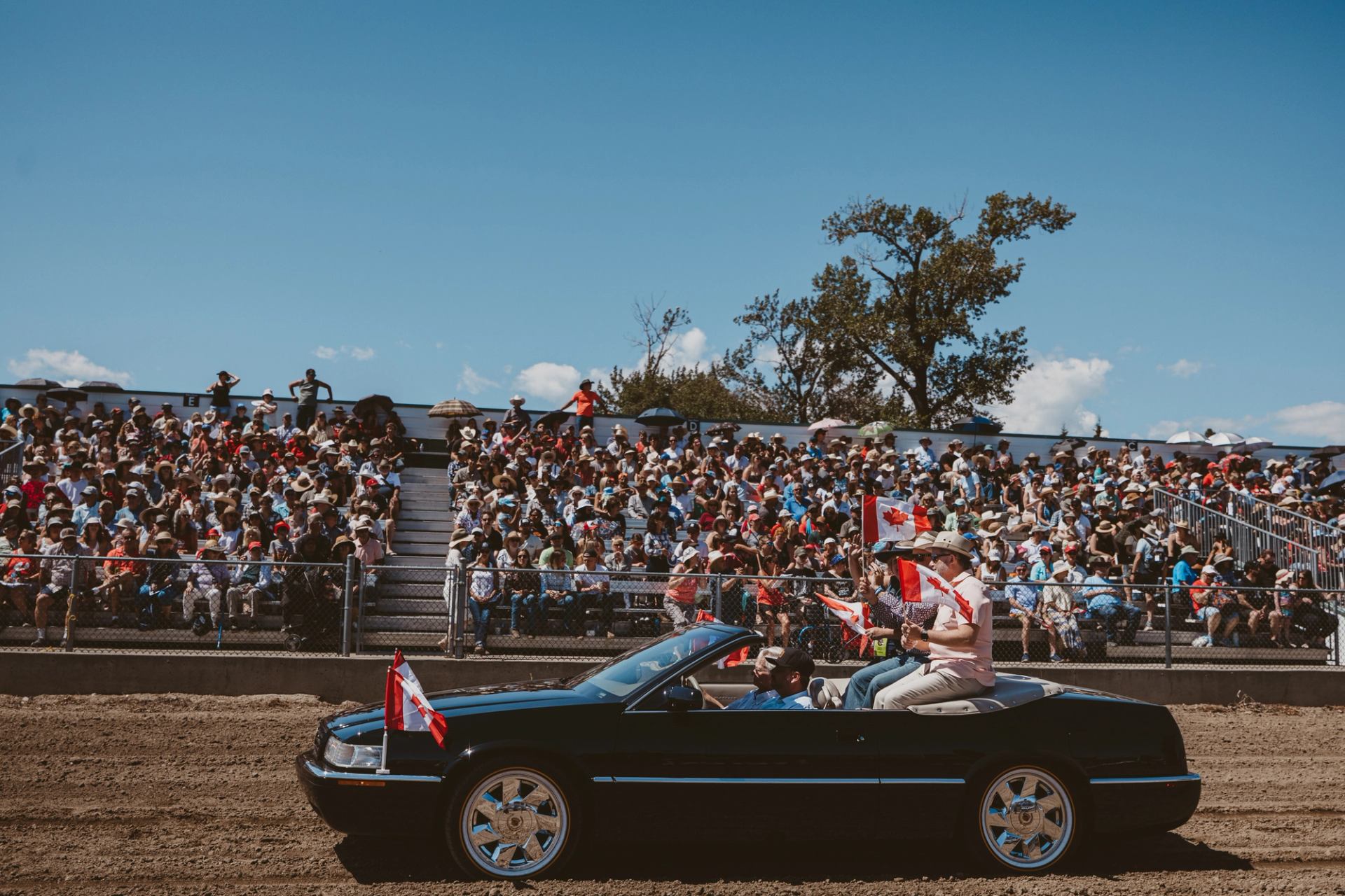 Convertible car drives along a dirt track with Canadian flags as grandstands filled with spectators look on.