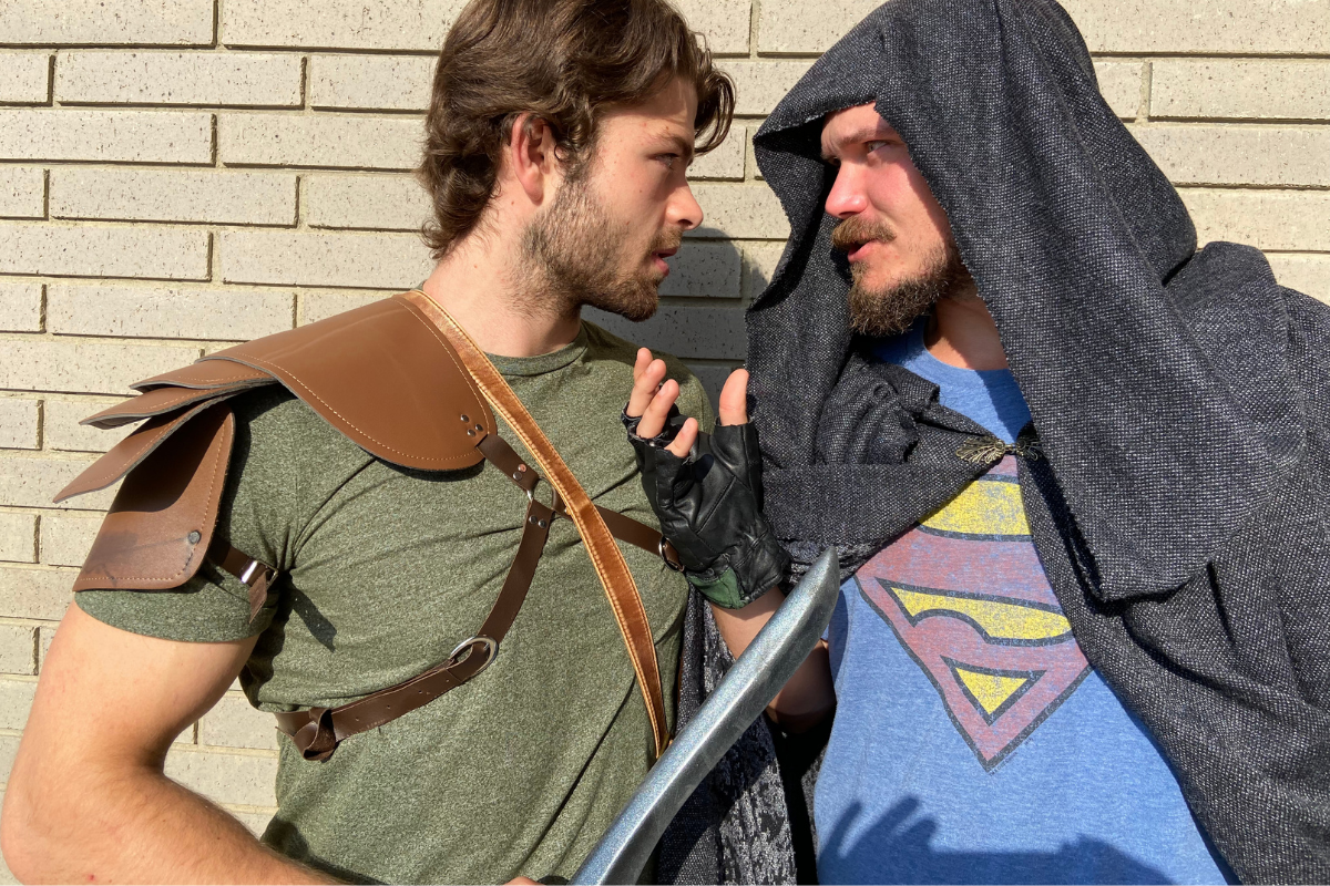 Two costumed people face each other, one in leather armour, one in hooded cloak.