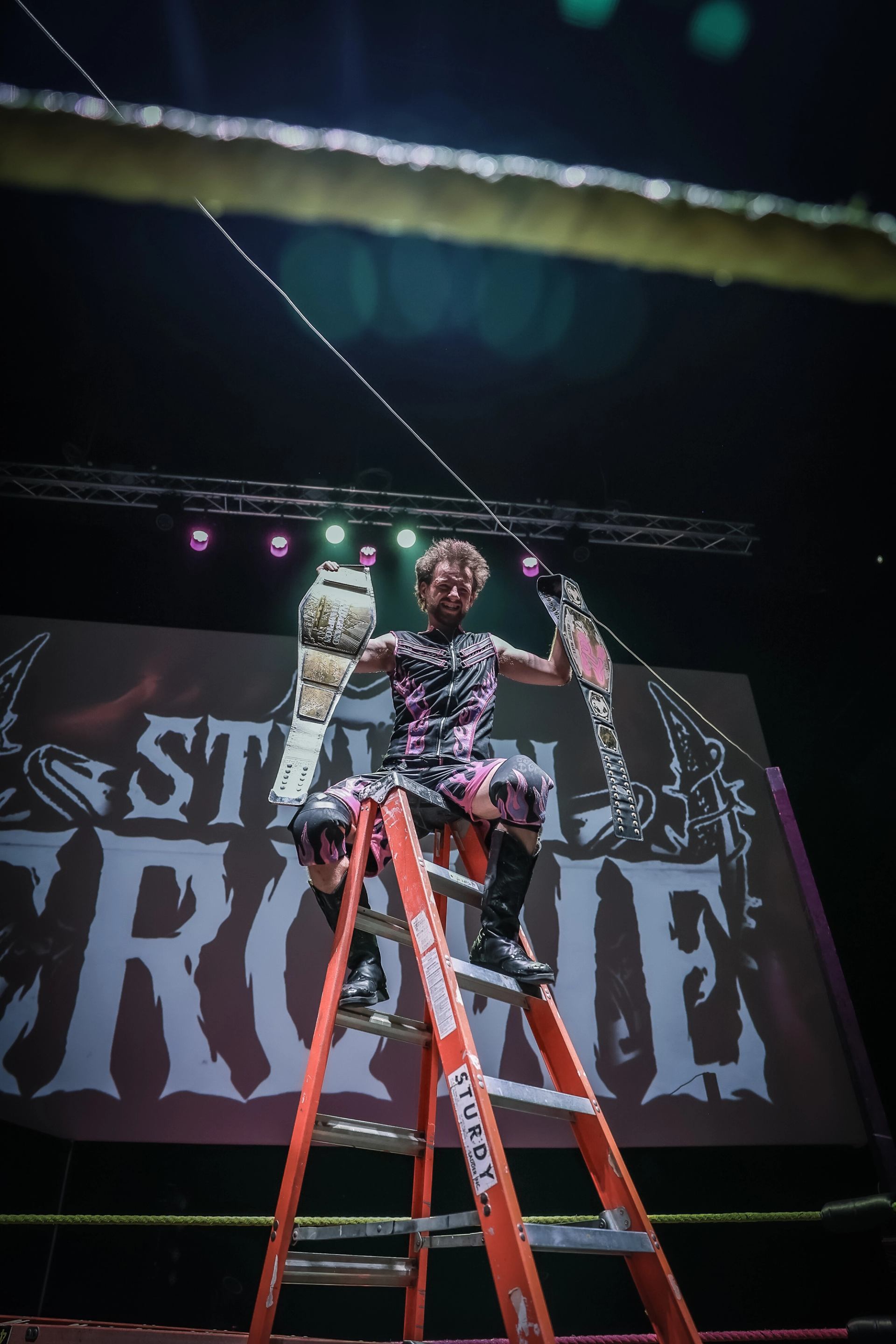 Wrestler standing atop a ladder holding two championship belts inside the ring at a Love Pro Wrestling show.