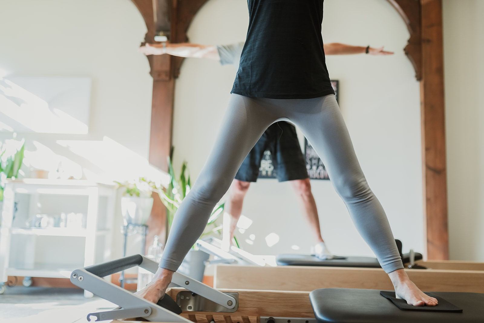 Woman performs pilates on a reformer, standing with hands on the platform indoors.