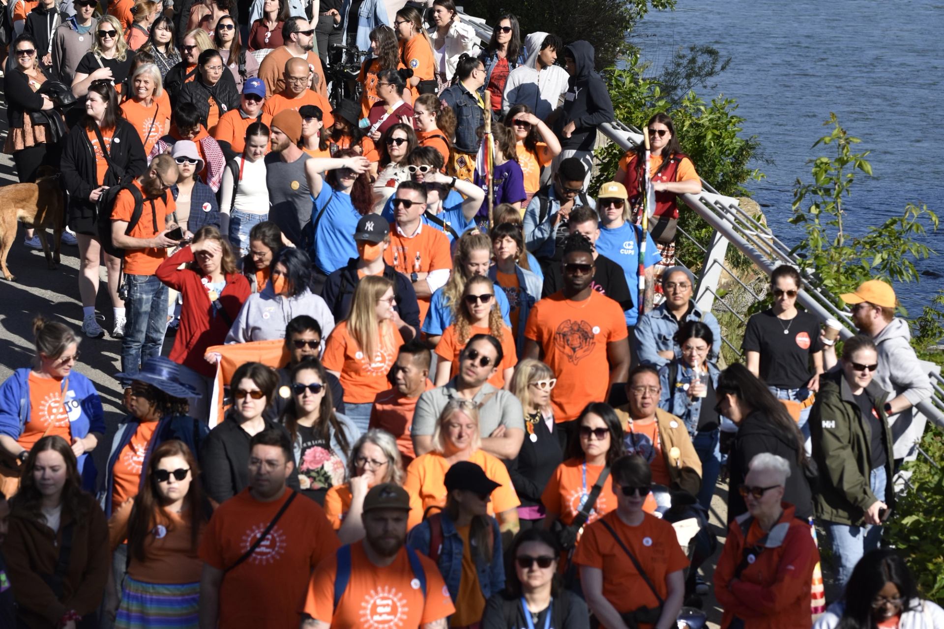 Large crowd wearing orange shirts walks along riverside path beside metal railing and trees.