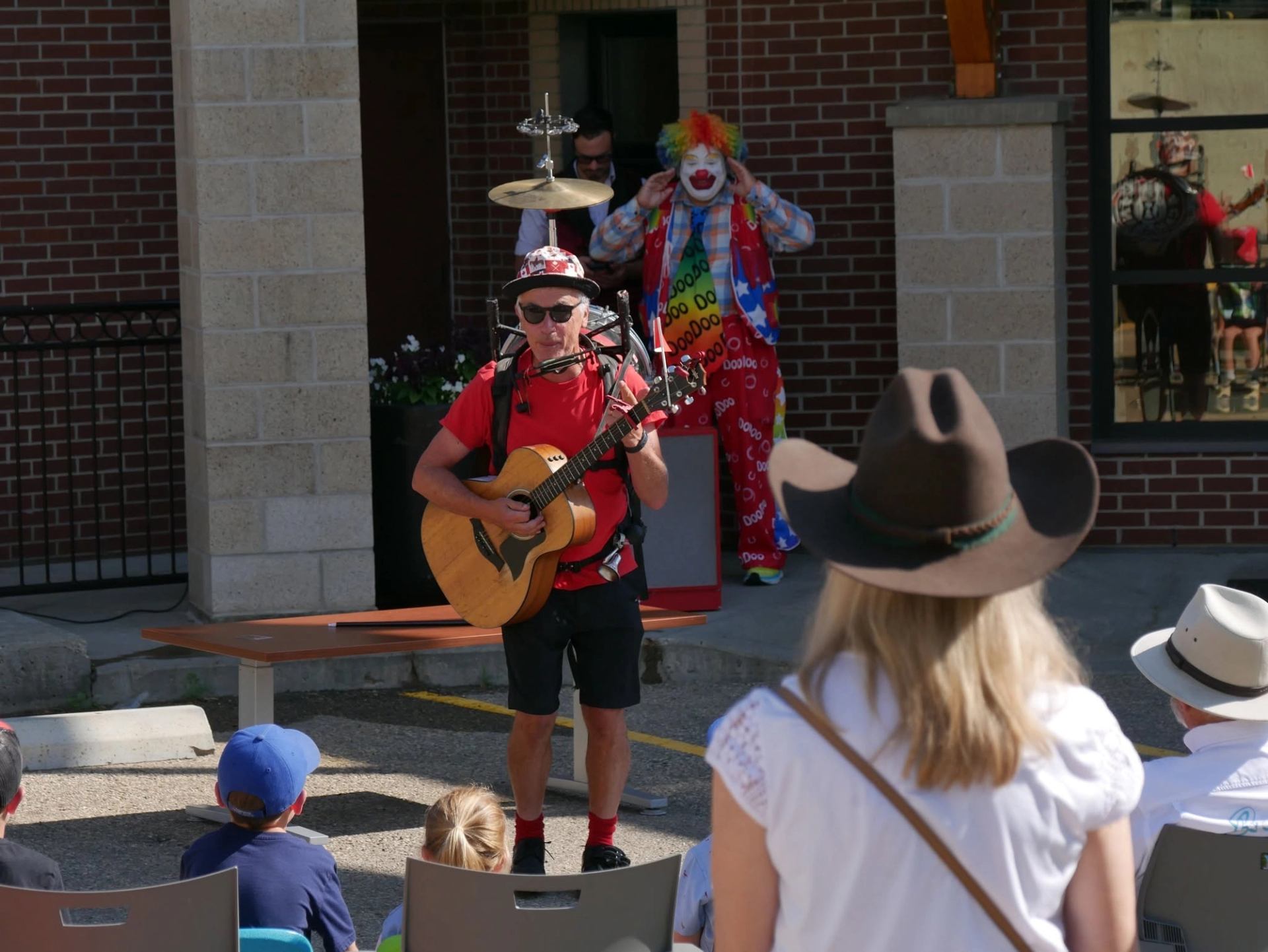 Street performer plays guitar and harmonica before a seated crowd as a clown stands behind.