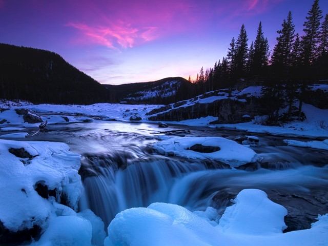 Snow-covered waterfall with purple sunset sky and pine trees in the distance