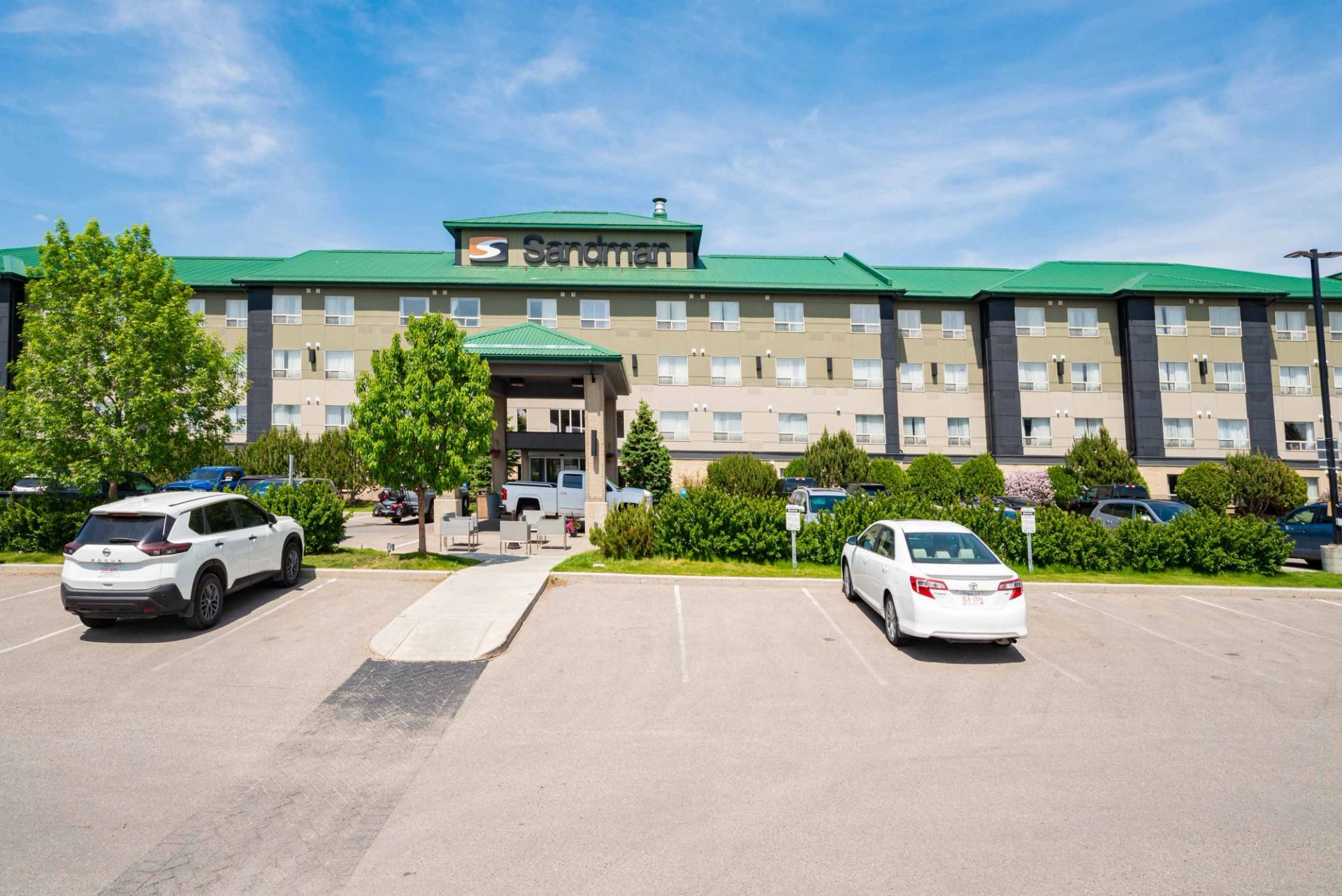 Sandman hotel exterior with green roof, front entrance, parking lot and parked cars.