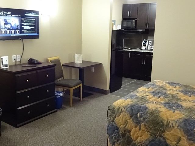 Hotel room with bed, desk, chair, kitchenette, and flat-screen TV mounted on wall.