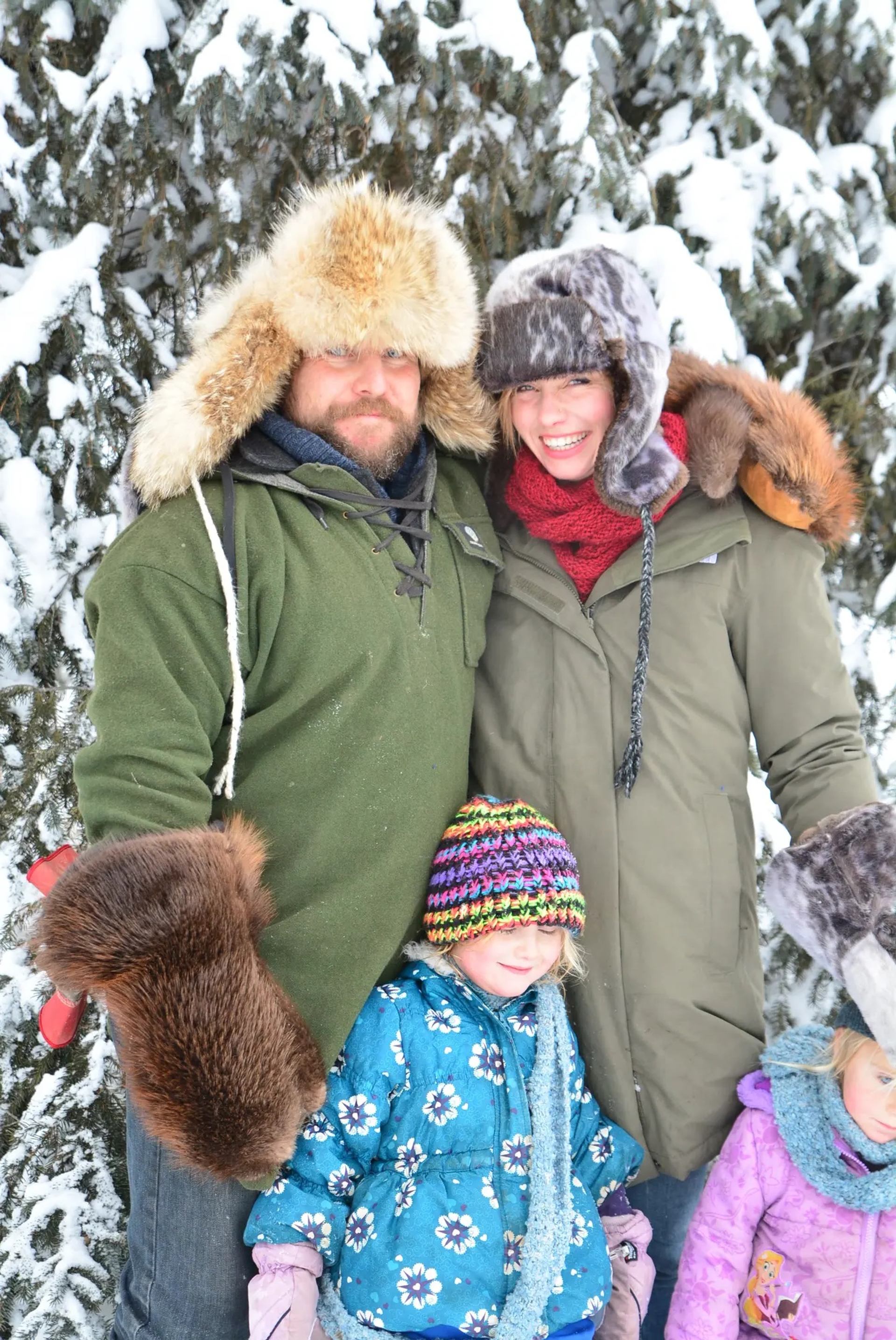 Family in winter clothes and fur hats smiling outdoors in snowy forest with evergreen trees.