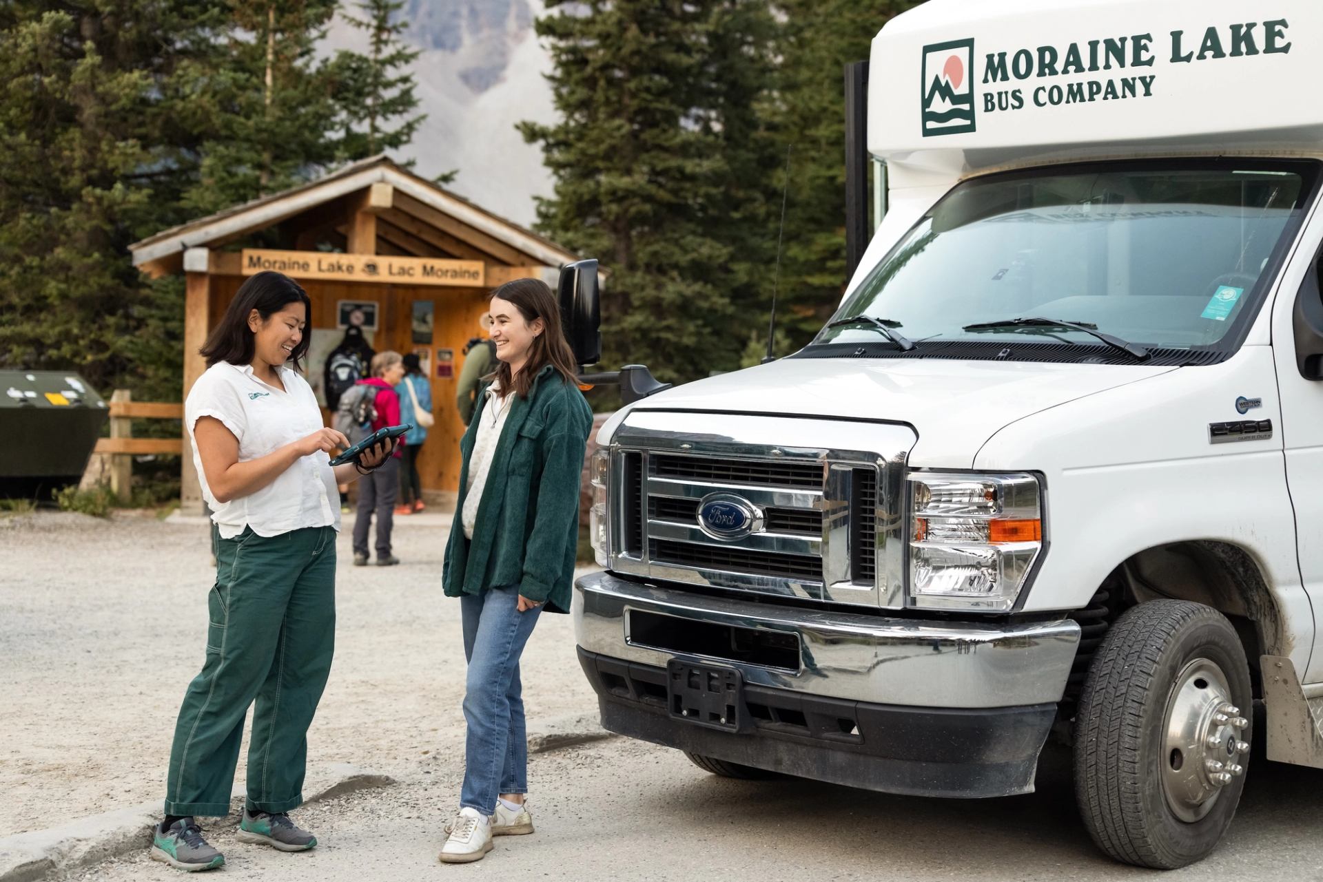 Staff helps visitor at Moraine Lake shuttle stop with bus and cabin in background.