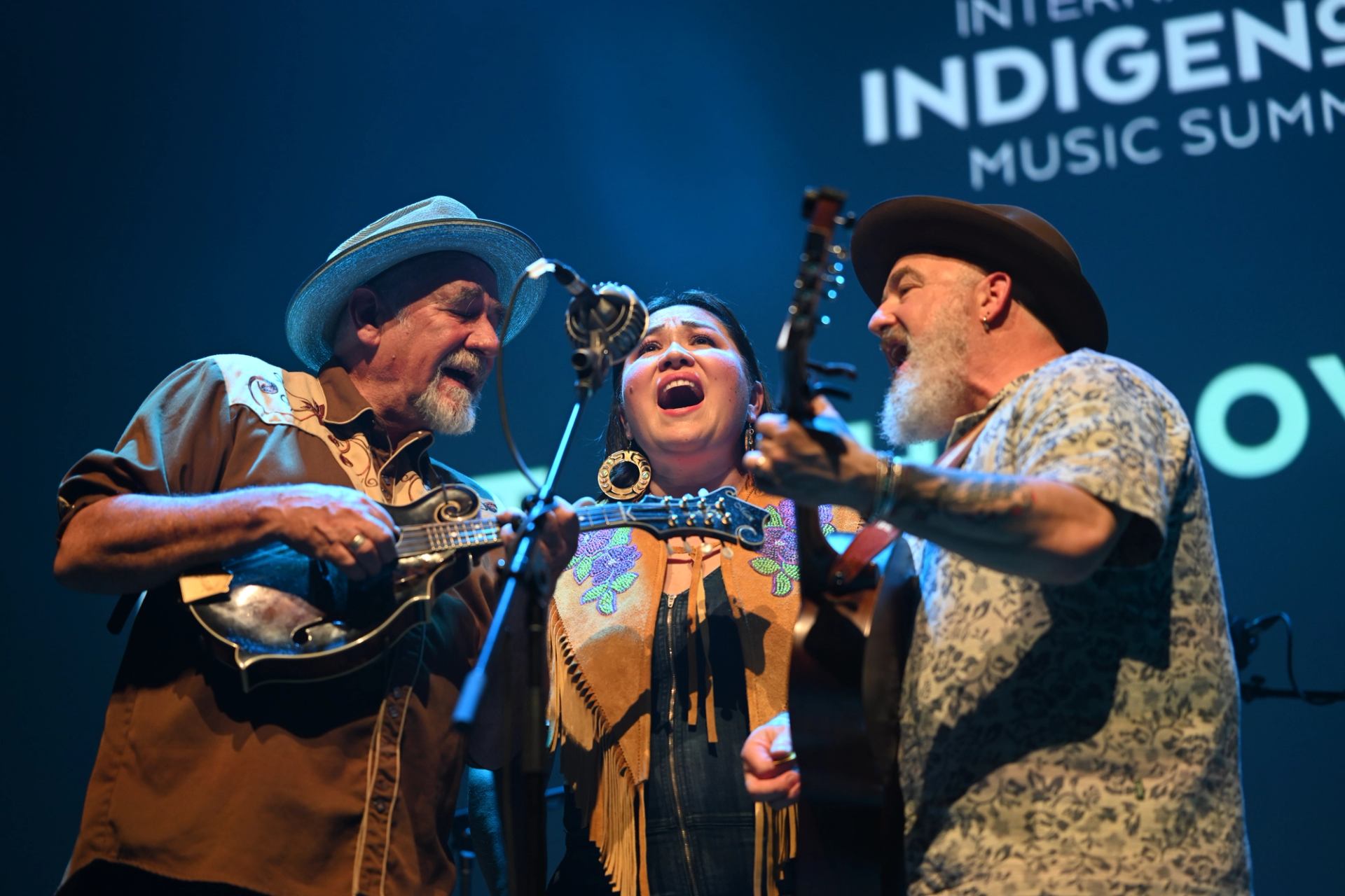 Three musicians sing into shared microphones while playing string instruments onstage.