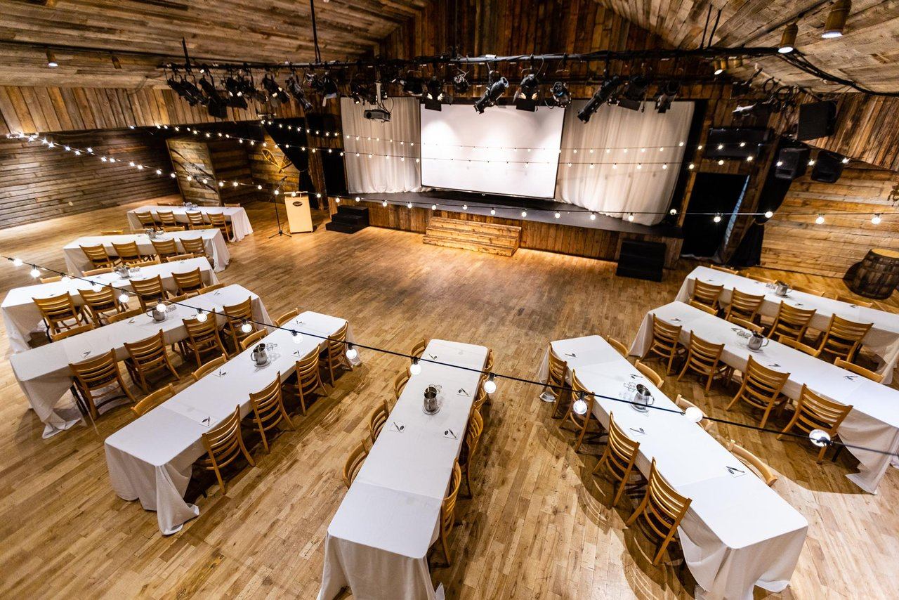 Large theatre hall with long white‑clothed tables and a wooden stage under string lights.