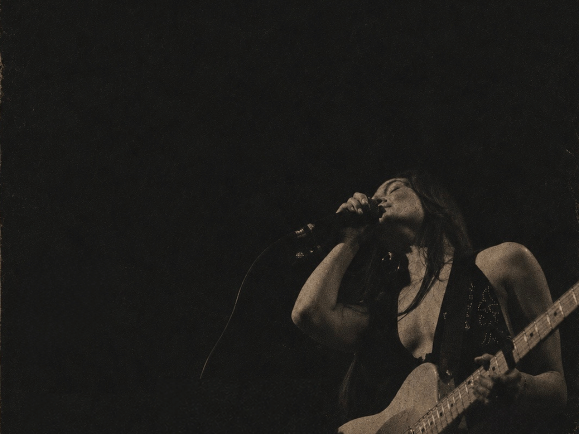 Singer holding electric guitar performs into microphone on dark, grainy stage background.