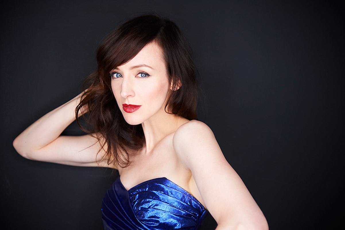 Woman in blue strapless dress posing against dark background, hand behind head.