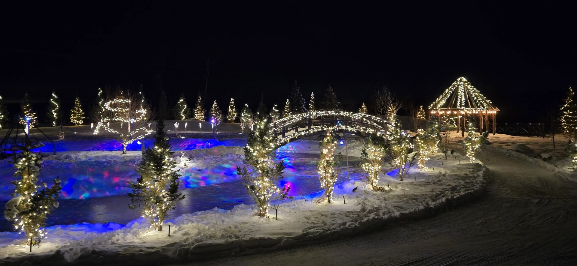 Snowy garden with glowing trees, bridges, and a lit gazebo at night.