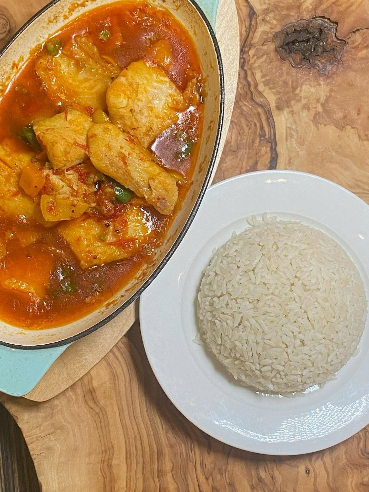 A rich tomato-based fish stew is served beside a dome of white rice on a wooden tabletop.